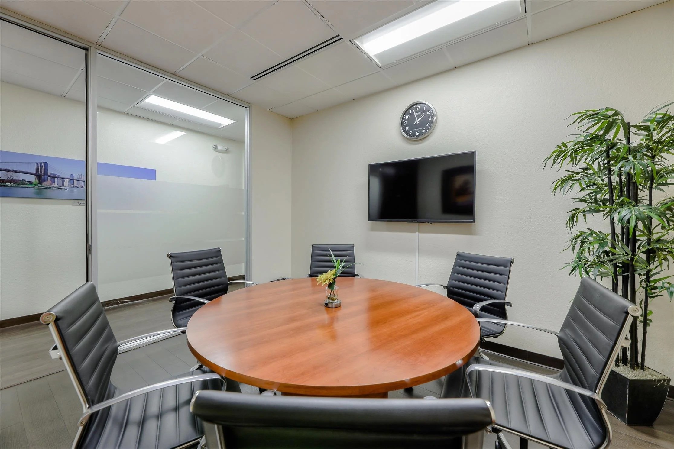Conference room with a wooden round table, six black leather chairs, a glass vase with a yellow flower, a large flat screen TV on the wall, a wall clock showing approximately 11:55, a potted plant, and a glass partition with a cityscape picture