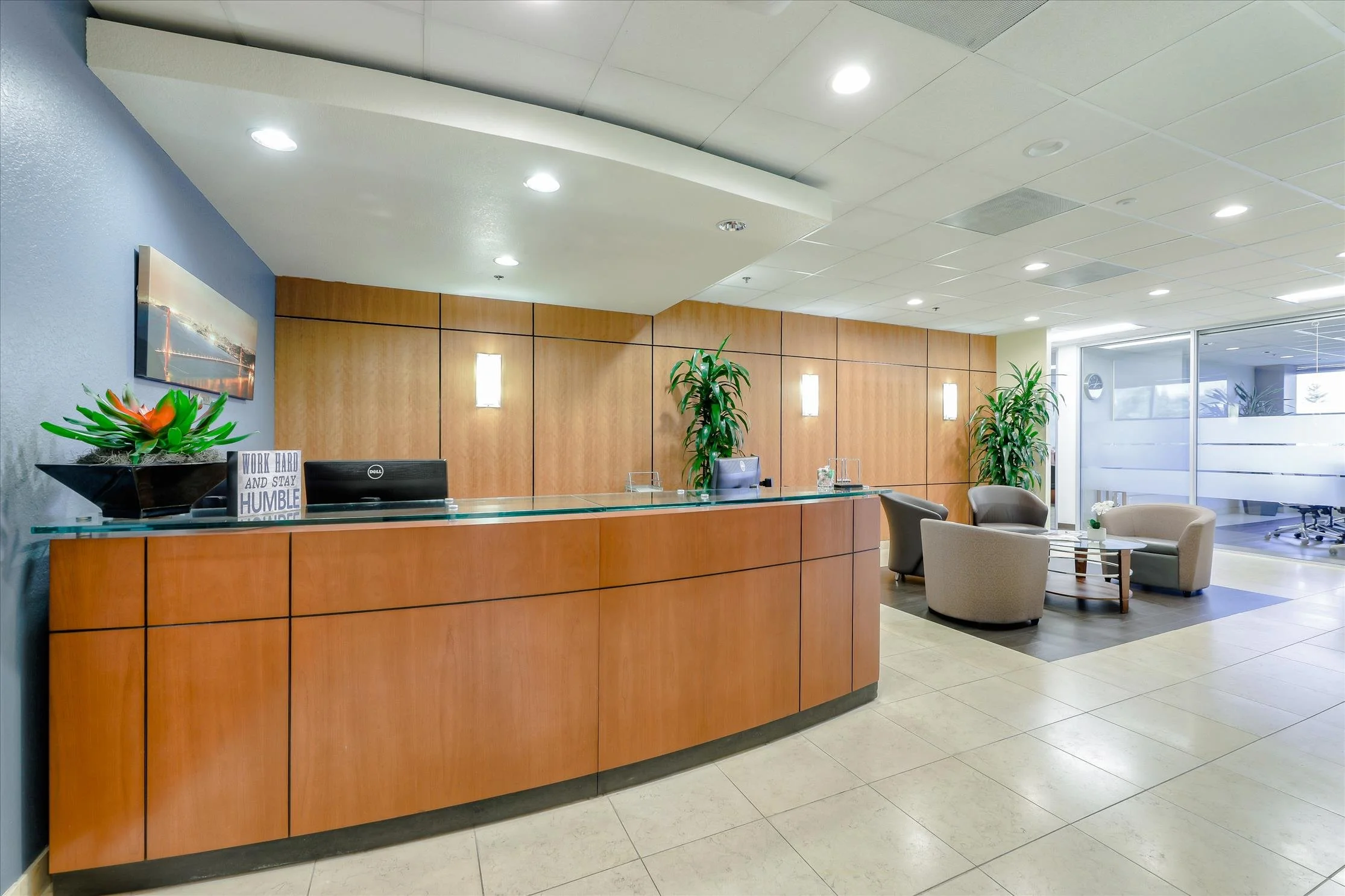 An office lobby with a wooden reception desk, potted plants, seating area with armchairs, and a glass wall in the background.