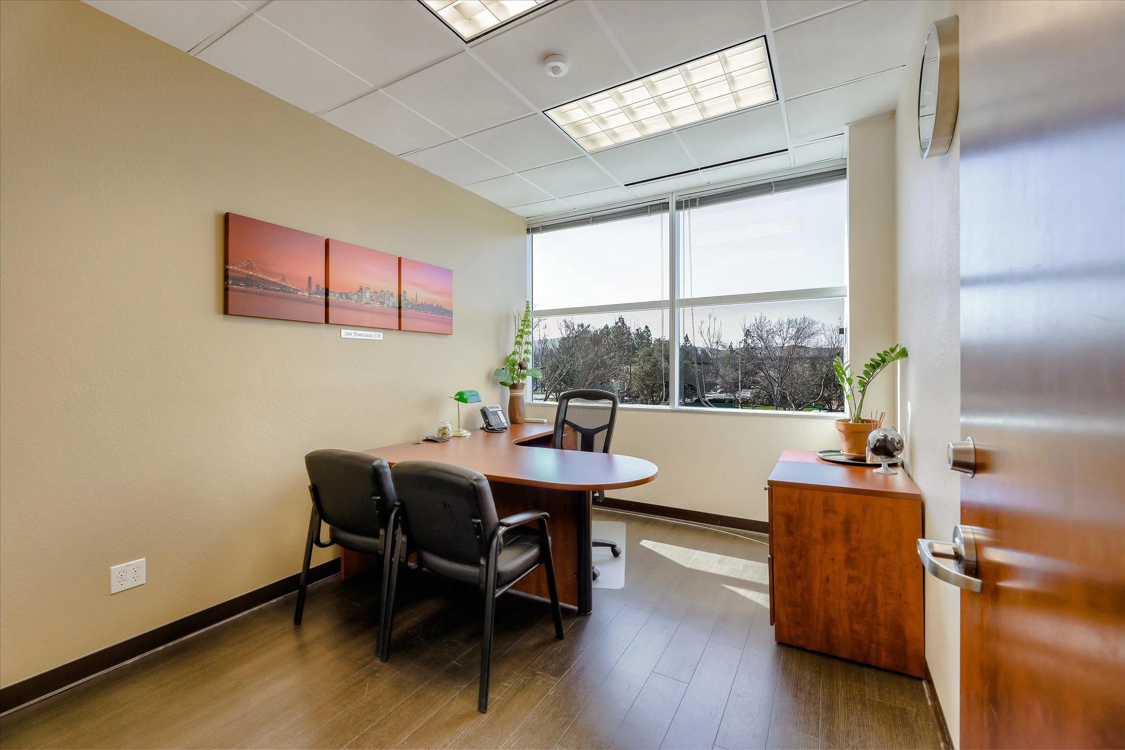 An office with beige walls, a large window showing trees, a wooden desk, black office chairs, and wall art of a cityscape with a bridge at sunset.