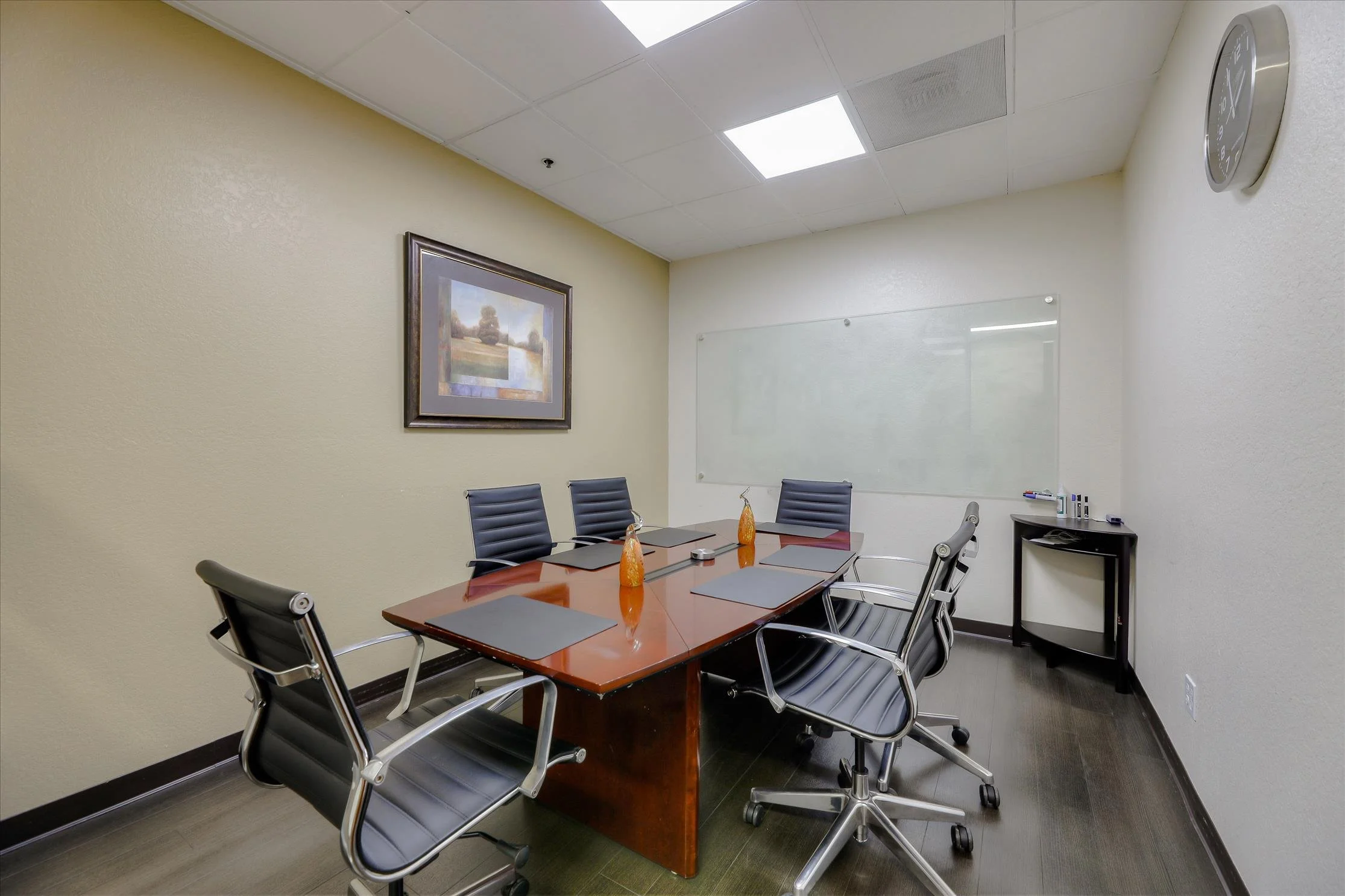 A conference room with a wooden table, six black leather chairs, a whiteboard, a framed landscape painting, a window, and a small black side table with markers and supplies.