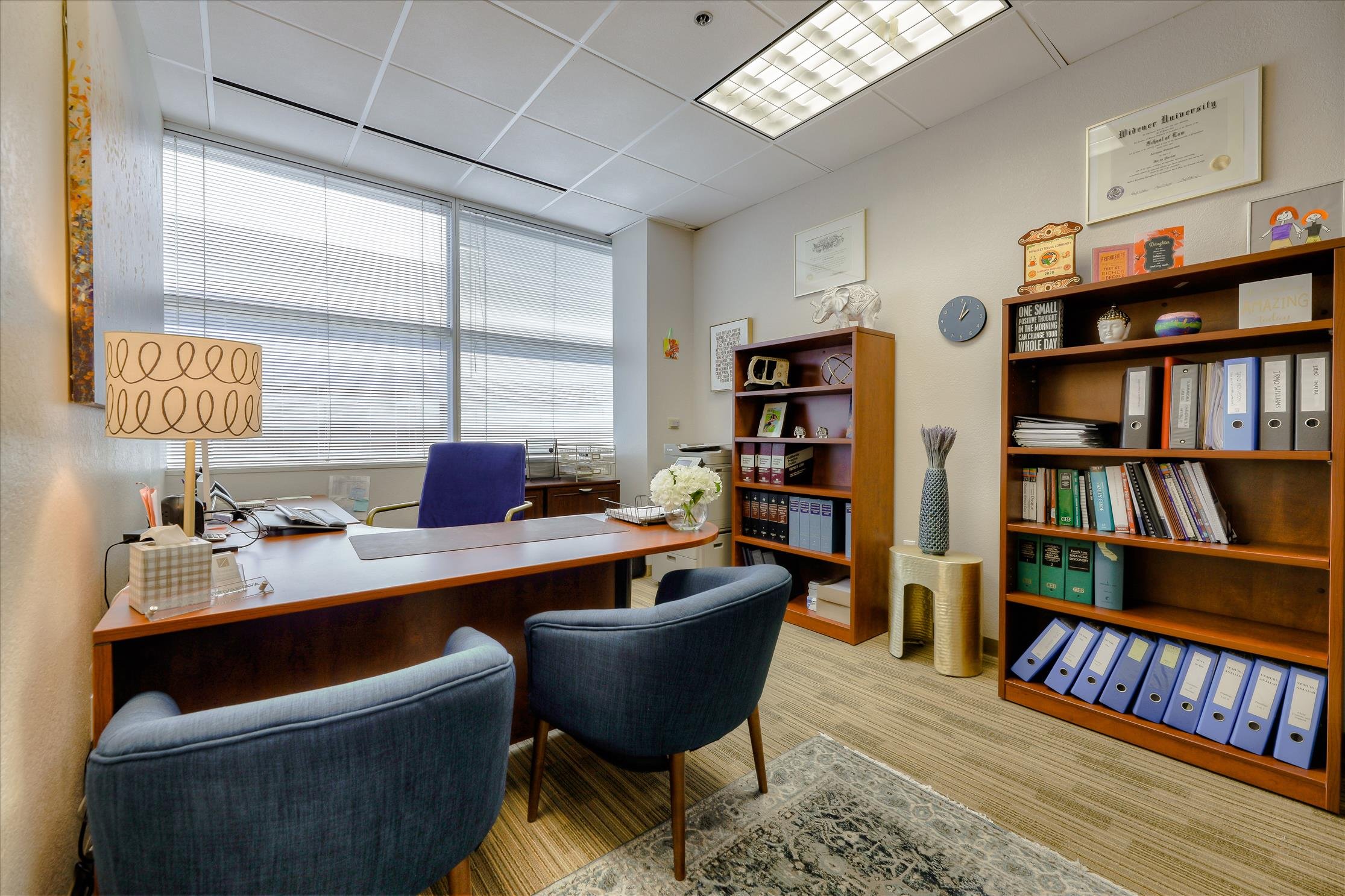 Office room with large windows, desk with office supplies, blue chairs, wooden bookshelves with binders and decorative objects, framed certificates on the wall, a ceiling with fluorescent lights.