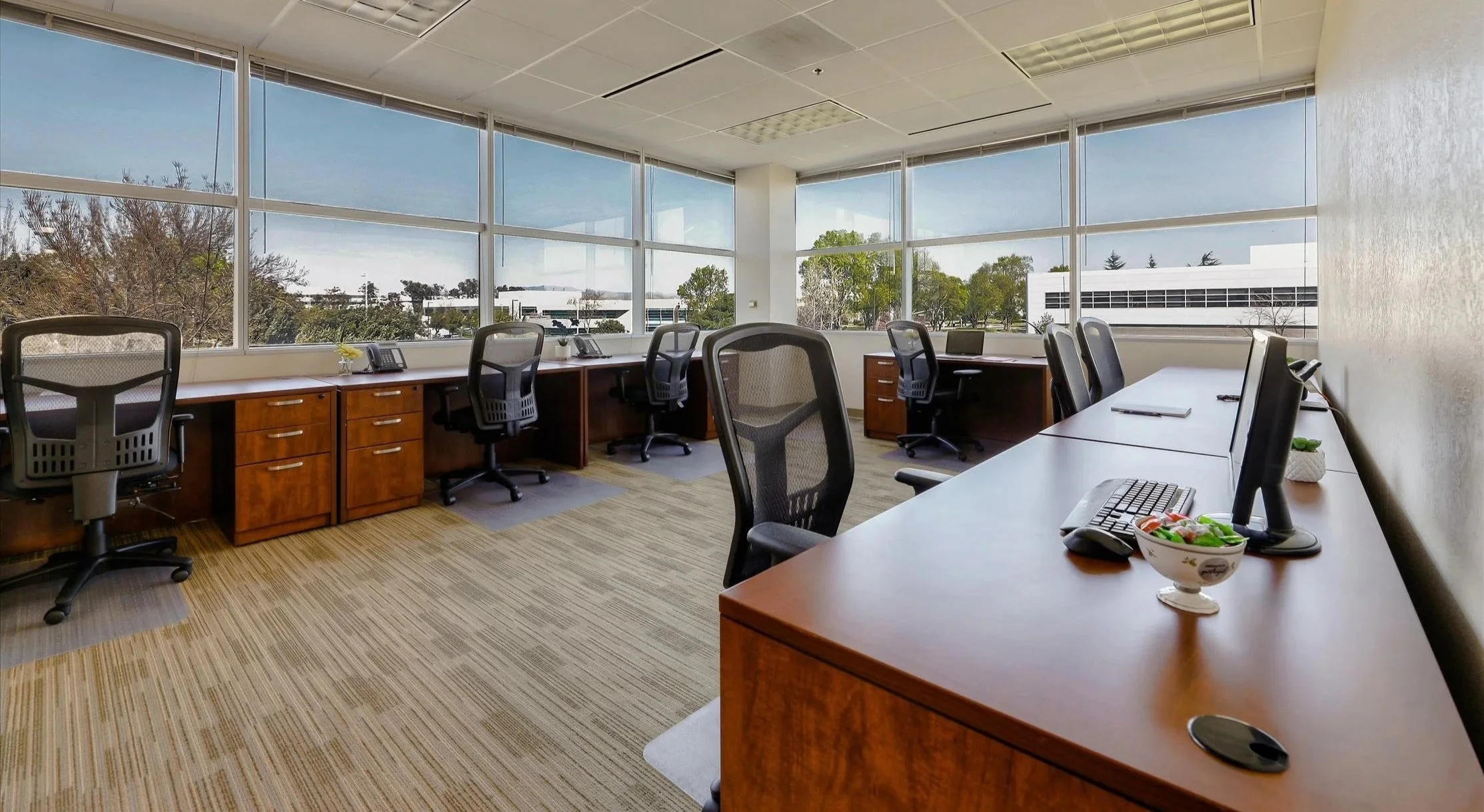 Office conference room with large windows, multiple desks with computer monitors, office chairs, and some personal items like a bowl of snacks and plants.