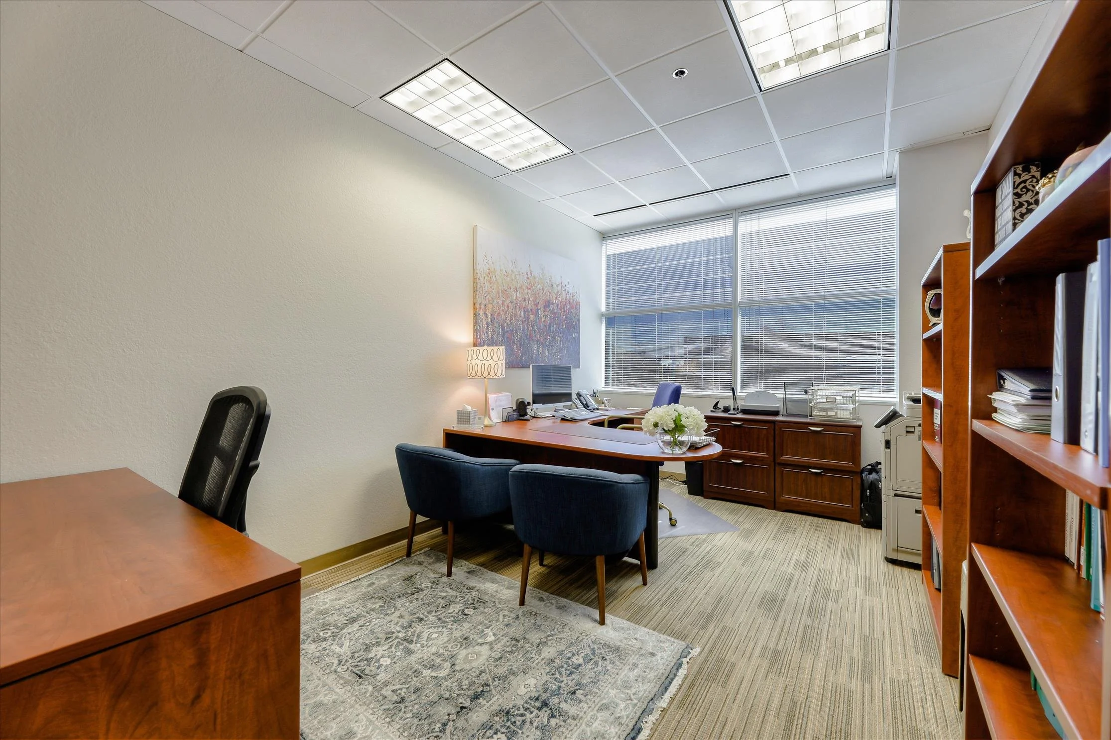 An office with a wooden desk, black office chair, and blue guest chairs. There are shelves with books, a printer, and a window with blinds letting in sunlight. A large painting hangs on the wall, and a lamp and vase with flowers are on the desk.