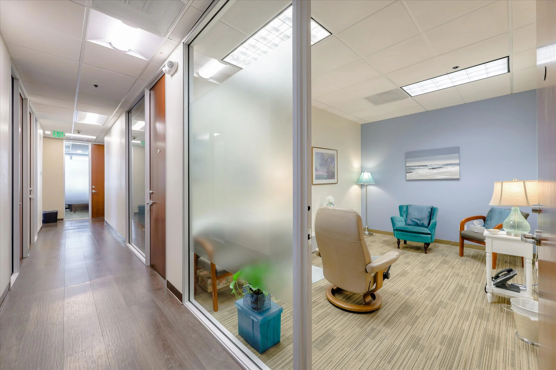 Office interior with hallway on the left, featuring doors and glass partitions, leading to a waiting or lounge area on the right with chairs, a floor lamp, artwork on the walls, and ceiling lights.