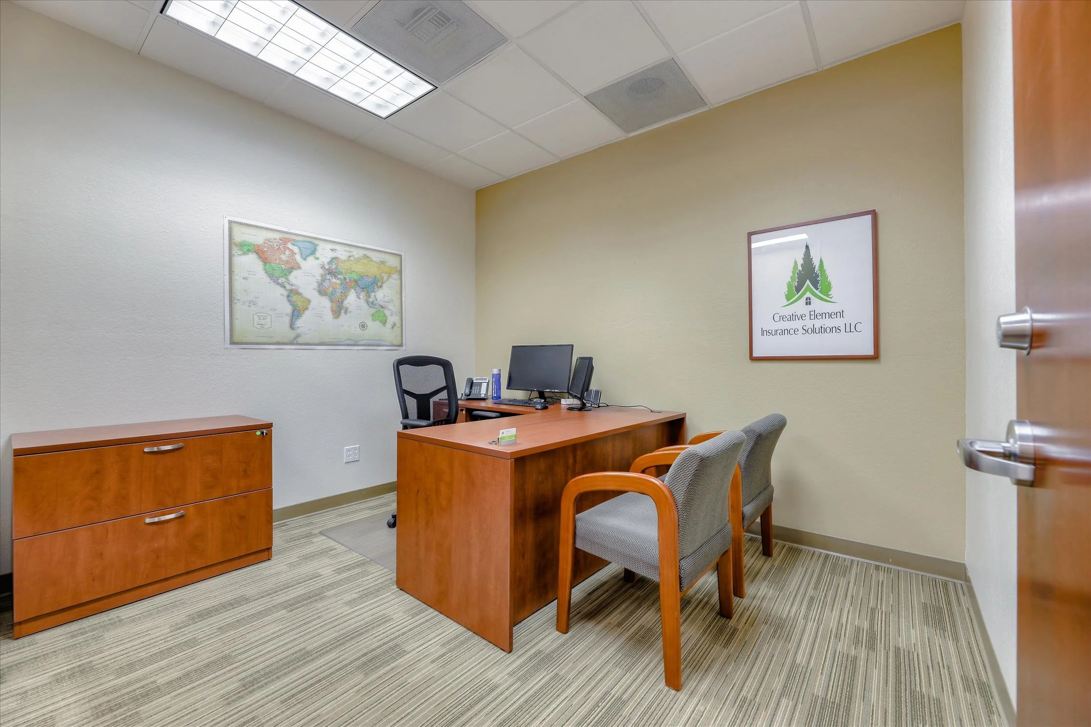 Office with a wooden desk and chairs, artwork on the wall, and a ceiling with fluorescent lights.