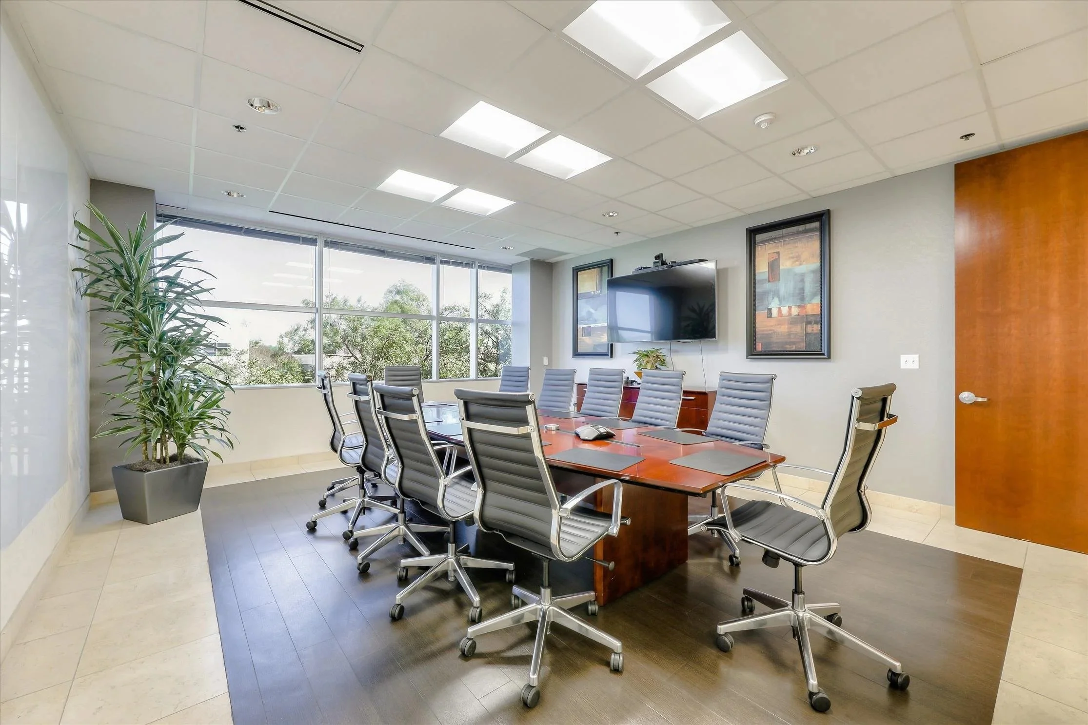 A modern conference room with a large wooden table surrounded by gray office chairs, a large flat-screen TV on the wall, framed artwork, a potted plant, and windows letting in natural light.