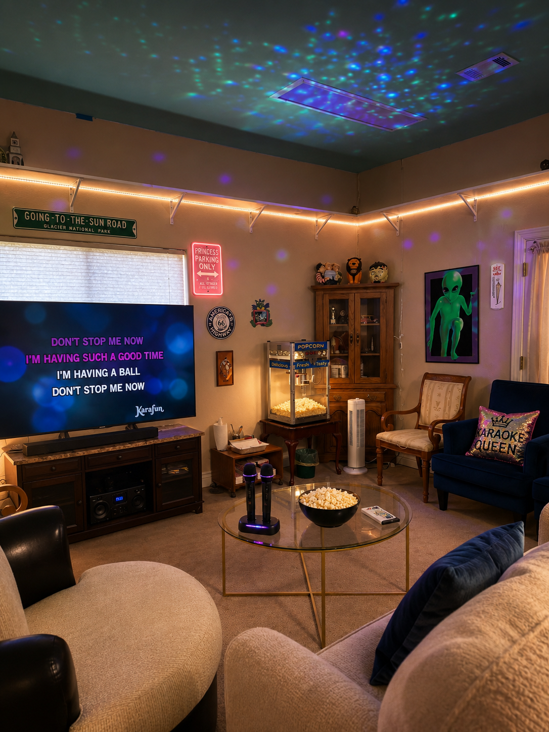 Living room with TV displaying karaoke lyrics, popcorn on a glass table, popcorn machine, and colorful ceiling lights. Furniture includes a beige armchair, a dark blue armchair with a princess crown pillow, and a wooden armchair. Decor includes a green alien poster, stuffed animals on a cabinet, and various signs and decorations.