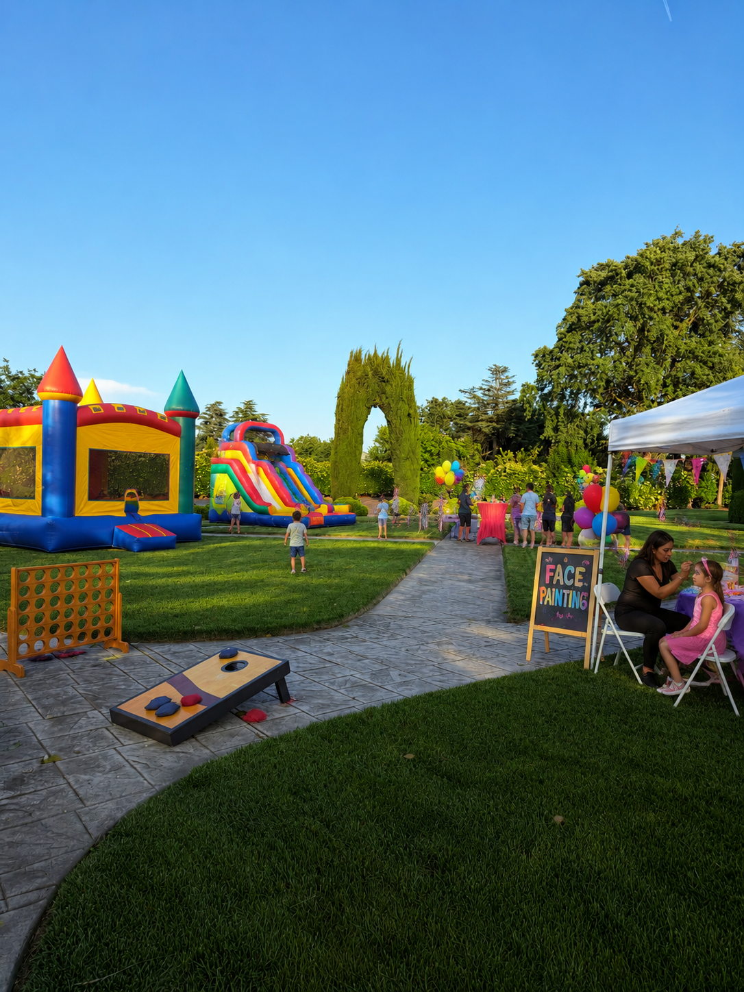Children playing on inflatable bounce house and slide at outdoor birthday party, face painting booth with woman and girl, decorated with colorful balloons and a sign that says 'Face Painting', on a sunny day in a lush green park.