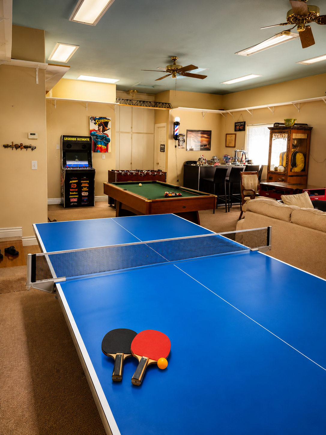 A basement game room with a blue ping pong table, a pool table, an arcade game, a foosball table, and various decorations and furniture.