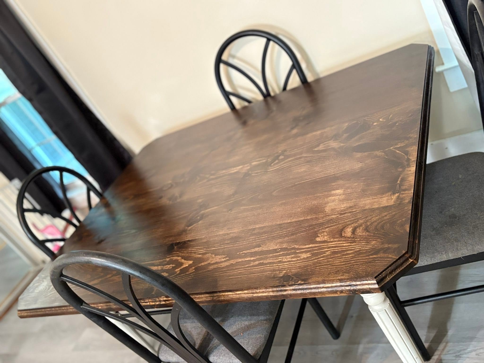 A wooden dining table with a dark brown finish, surrounded by black metal chairs with curved backs. The table is positioned in a room with beige walls, and the photograph is taken from a corner, showing part of a black frame mirror or picture frame reflecting onto the table.
