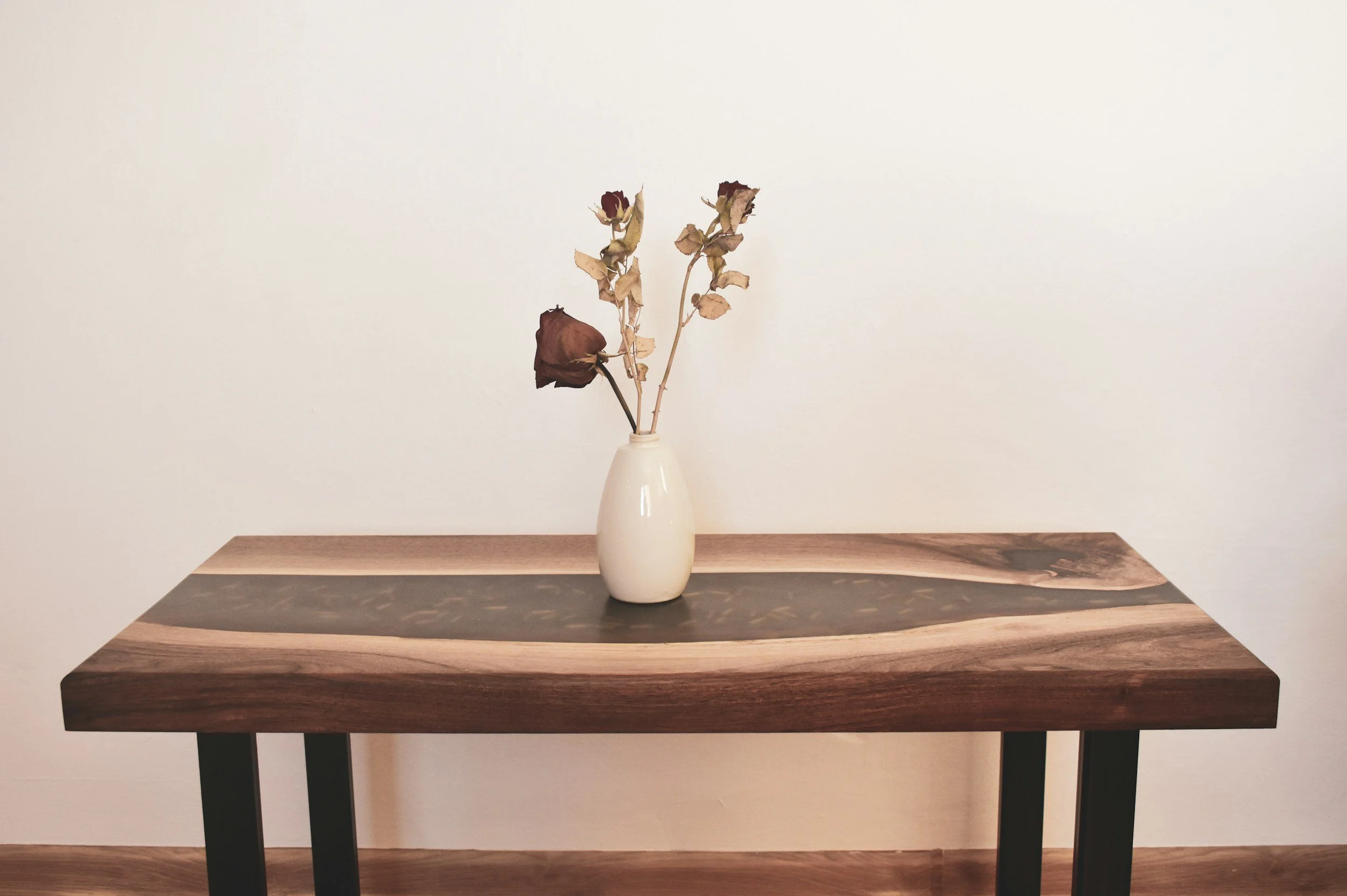 A white vase with dried roses, placed on a decorative wooden table with a two-tone wood pattern, against a plain white wall.
