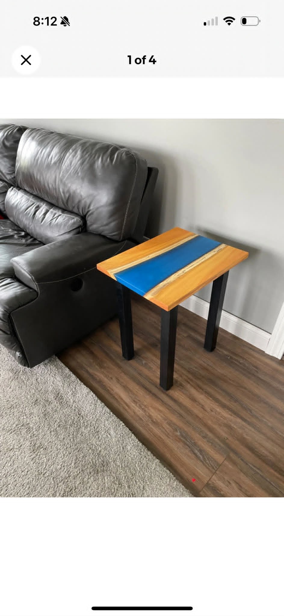 A black leather couch next to a small wooden side table with a blue and natural wood pattern top, on a wooden floor next to a gray wall.