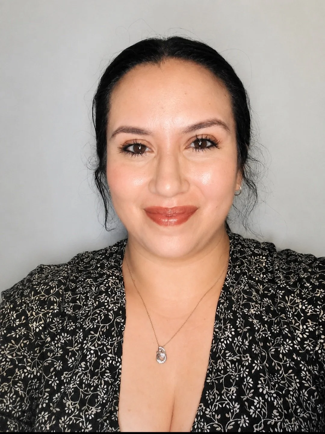 A woman with dark hair pulled back, wearing a black and white floral patterned top, gold earrings, and a necklace with a pendant, smiling at the camera in front of a plain gray background.