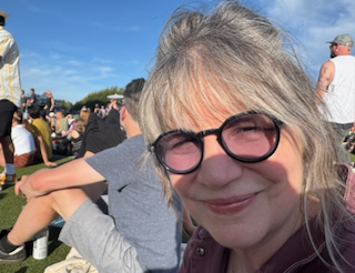A woman with gray hair and black glasses smiling outdoors at an event with a crowd and blue sky in the background.