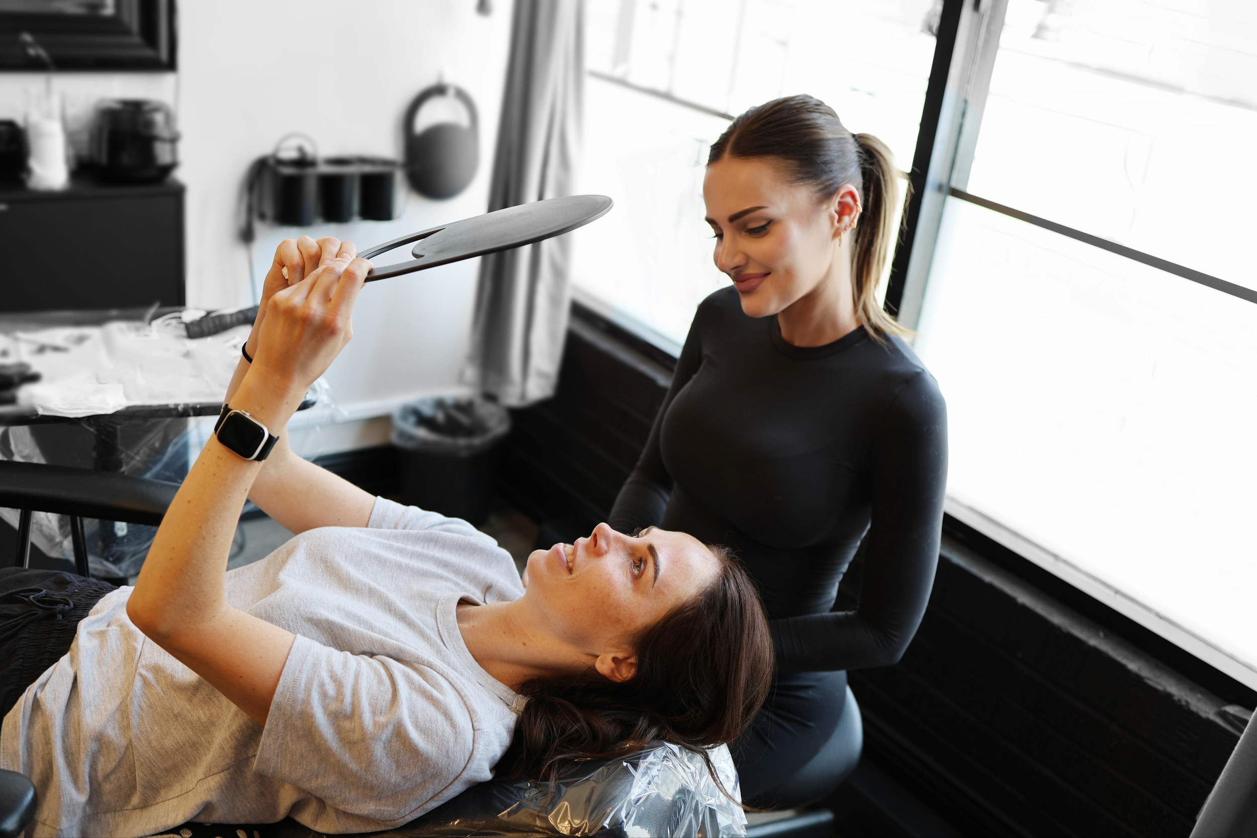 A woman lying down at a salon or spa, smiling, with her head on a cushion, receiving a manicure or beauty treatment from another woman who is holding a mirror and smiling, in a bright room with large windows.
