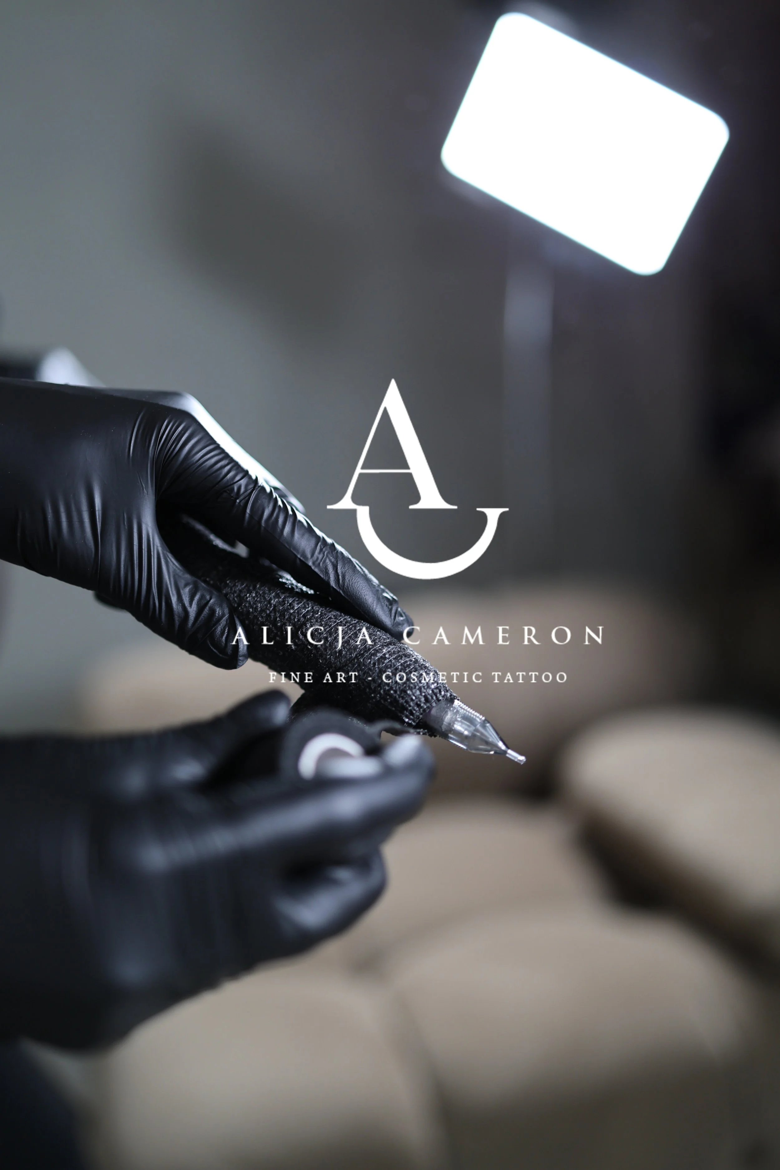 Close-up of a hand wearing black gloves holding a tattoo machine, with a bright light in the background. Text on the image: Alicia Cameron, Fine Art Cosmetic Tattoo.