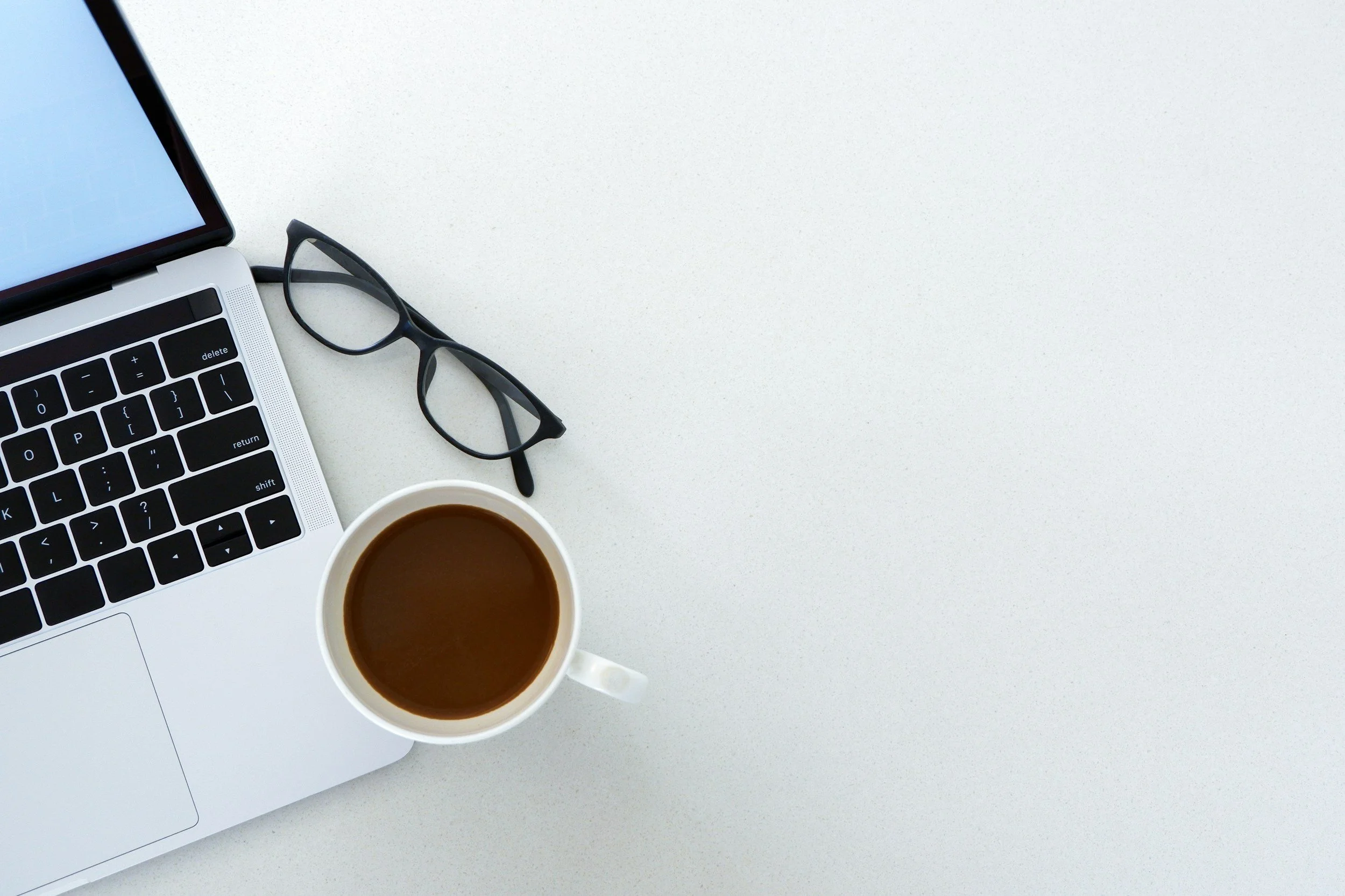 A white desk with a part of a silver laptop, a pair of black eyeglasses, a white coffee cup filled with coffee, and a portion of a smartphone.