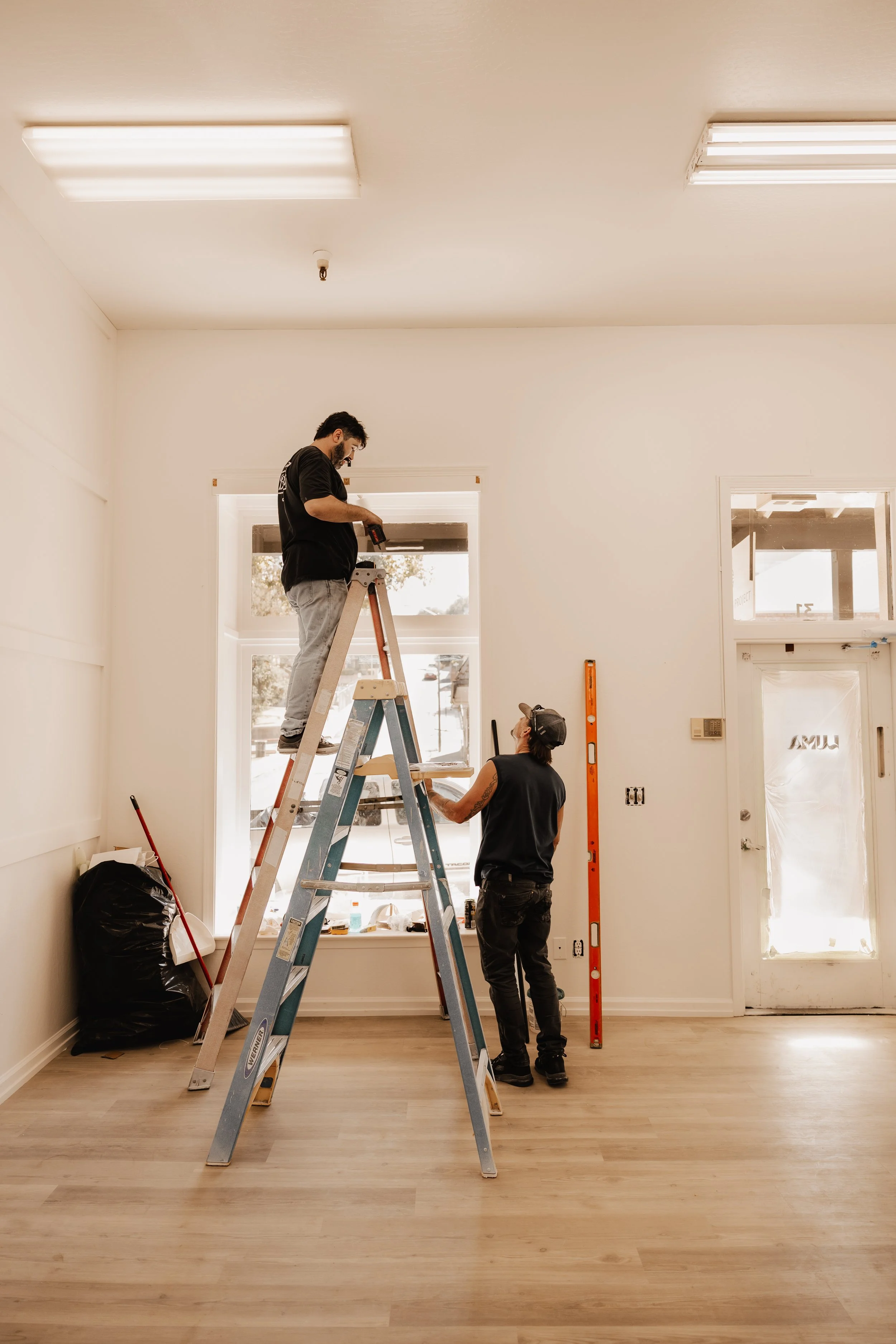 Two workers installing or repairing a window in a bright, empty room with light-colored wood flooring. One person is standing on a ladder and another is on the floor, both engaged in the task.