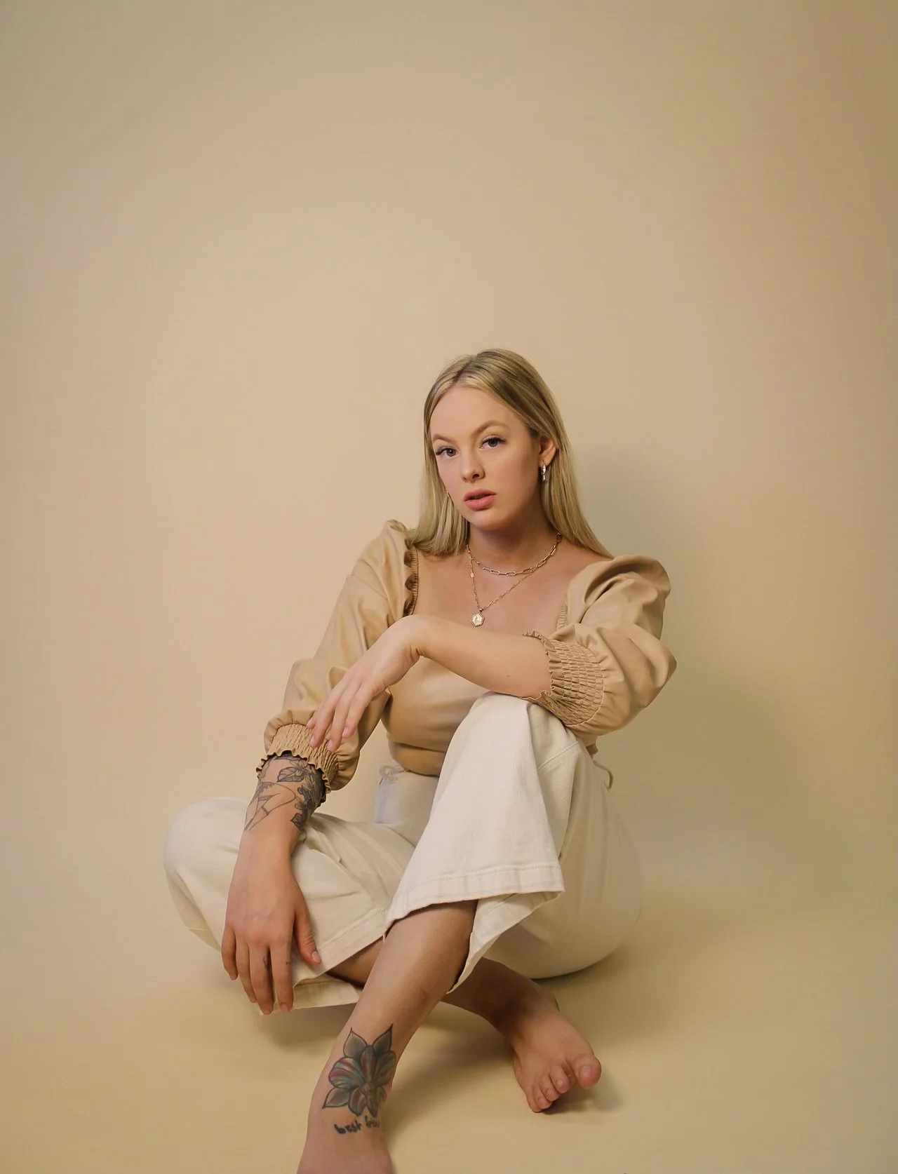 A young woman with blonde hair and tattoos sitting on the floor against a plain beige wall, wearing beige top and cream-colored pants, accessorized with silver jewelry.