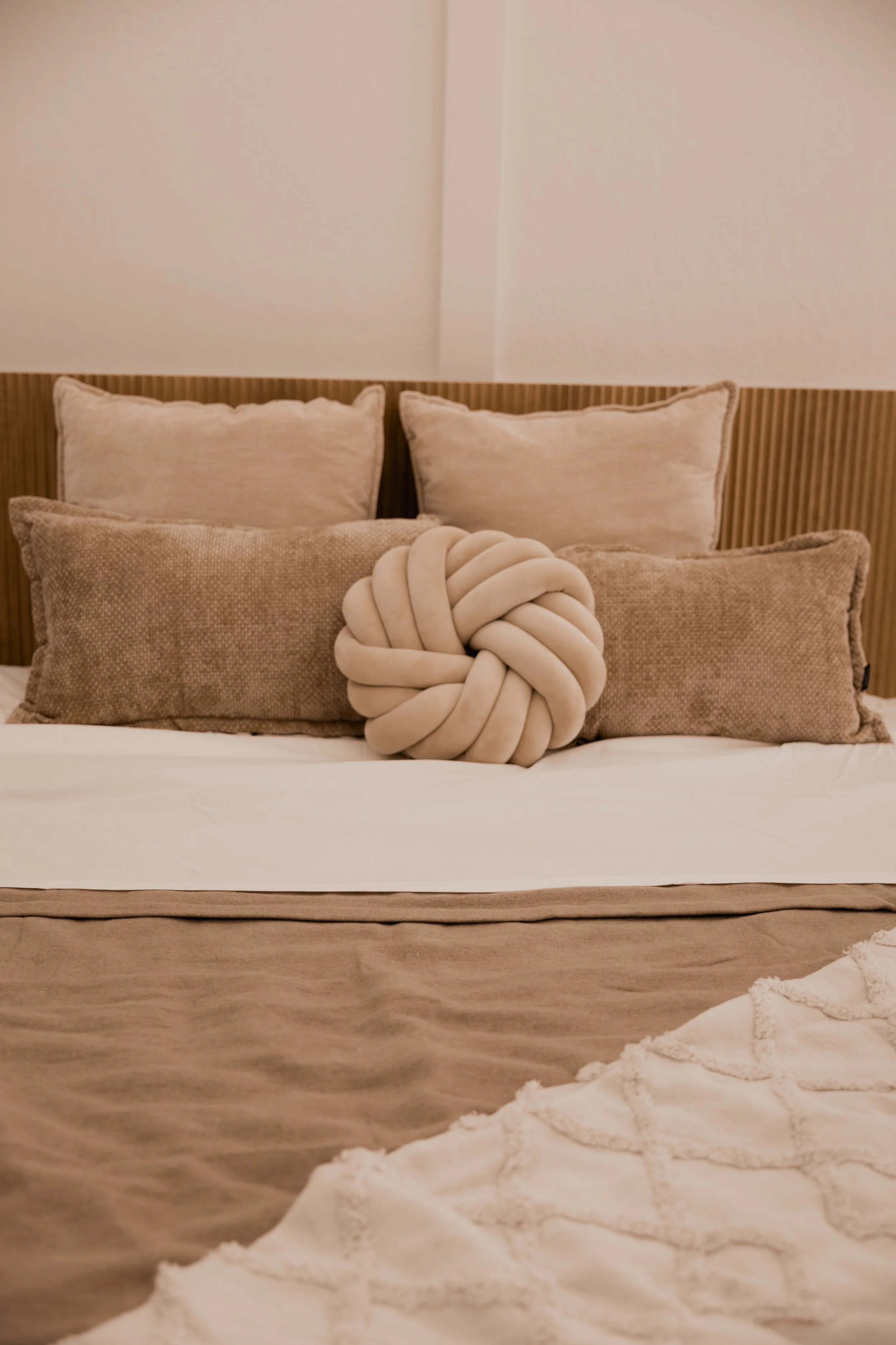 A neatly made bed with beige pillows, a decorative knot pillow, a textured square pillow, a padded headboard, and a beige wall behind.