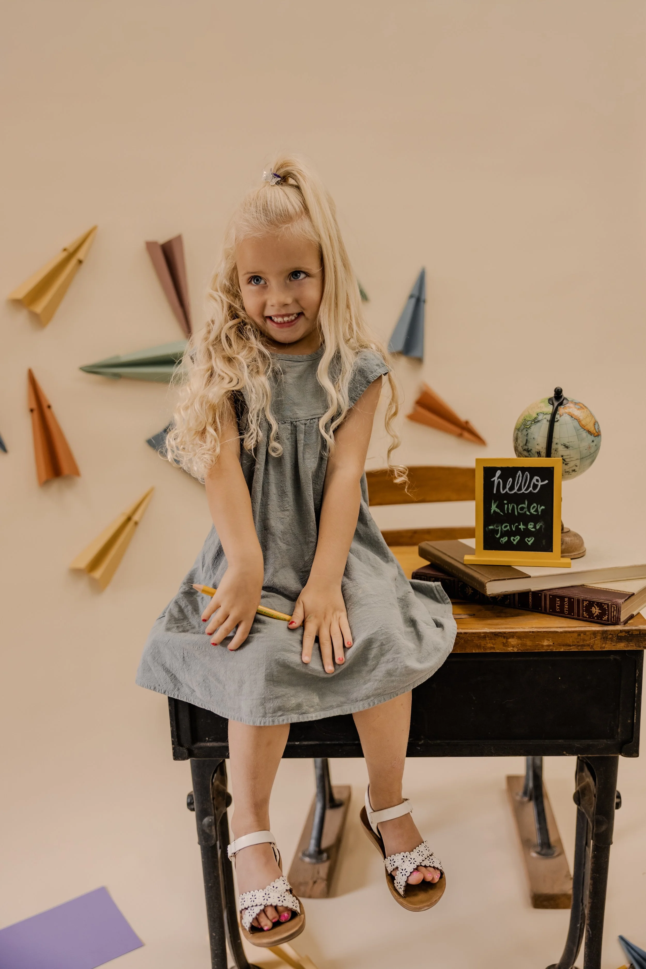 A young girl with long blonde hair sits on a vintage black sewing machine table, smiling. She wears a gray dress, white sandals, and has a small bow in her hair. Behind her are colorful paper airplanes on a beige wall, and a small chalkboard with the message 'hello kinder garden' and hearts, next to a globe.
