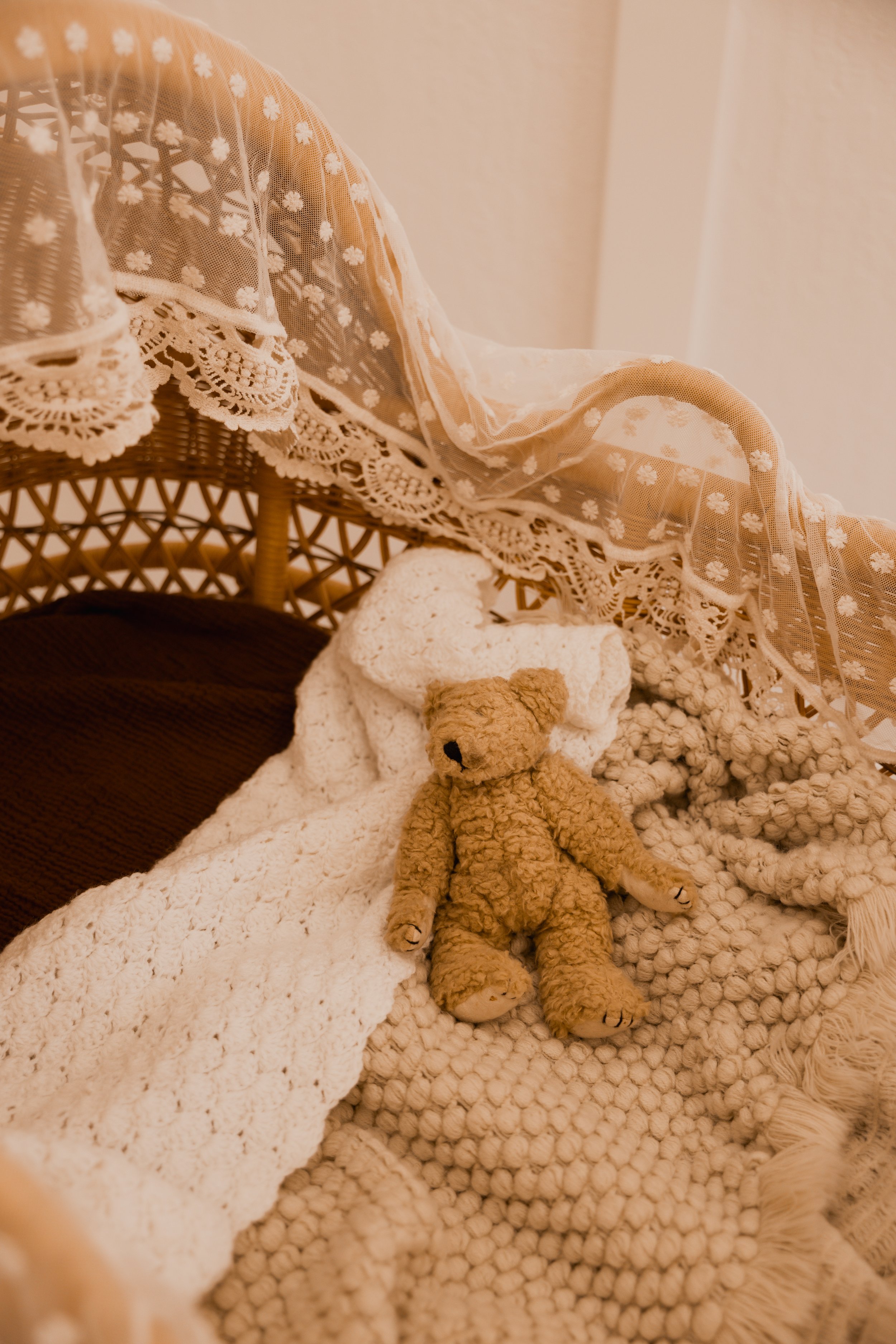 A close-up of a cozy, vintage-style scene with a teddy bear, textured blankets, a wicker chair with lace detailing, and sheer fabric with floral patterns.