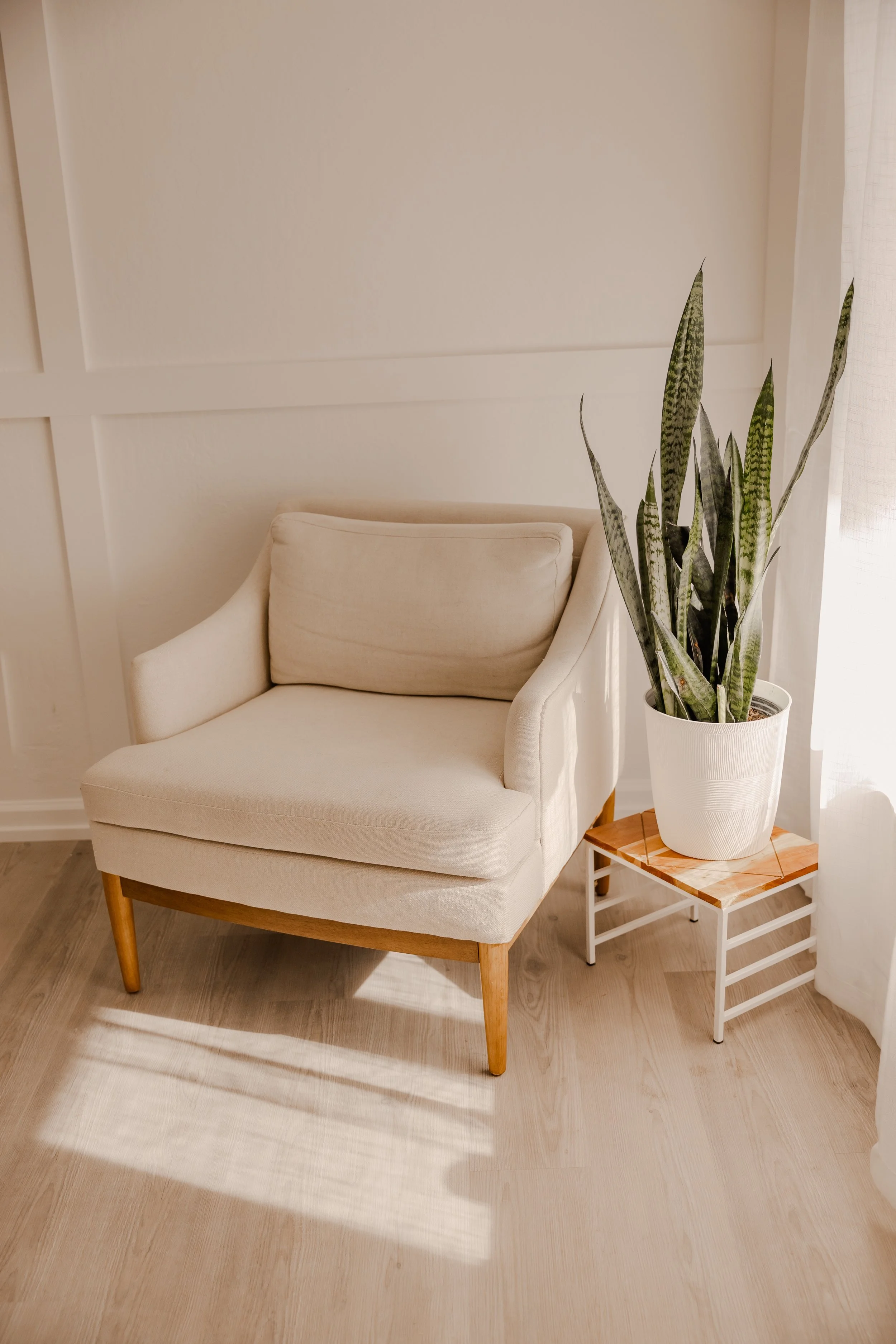 A beige armchair with wooden legs, next to a small white side table with a potted snake plant that has long, variegated green and gray leaves, in a room with light wooden flooring and white walls.
