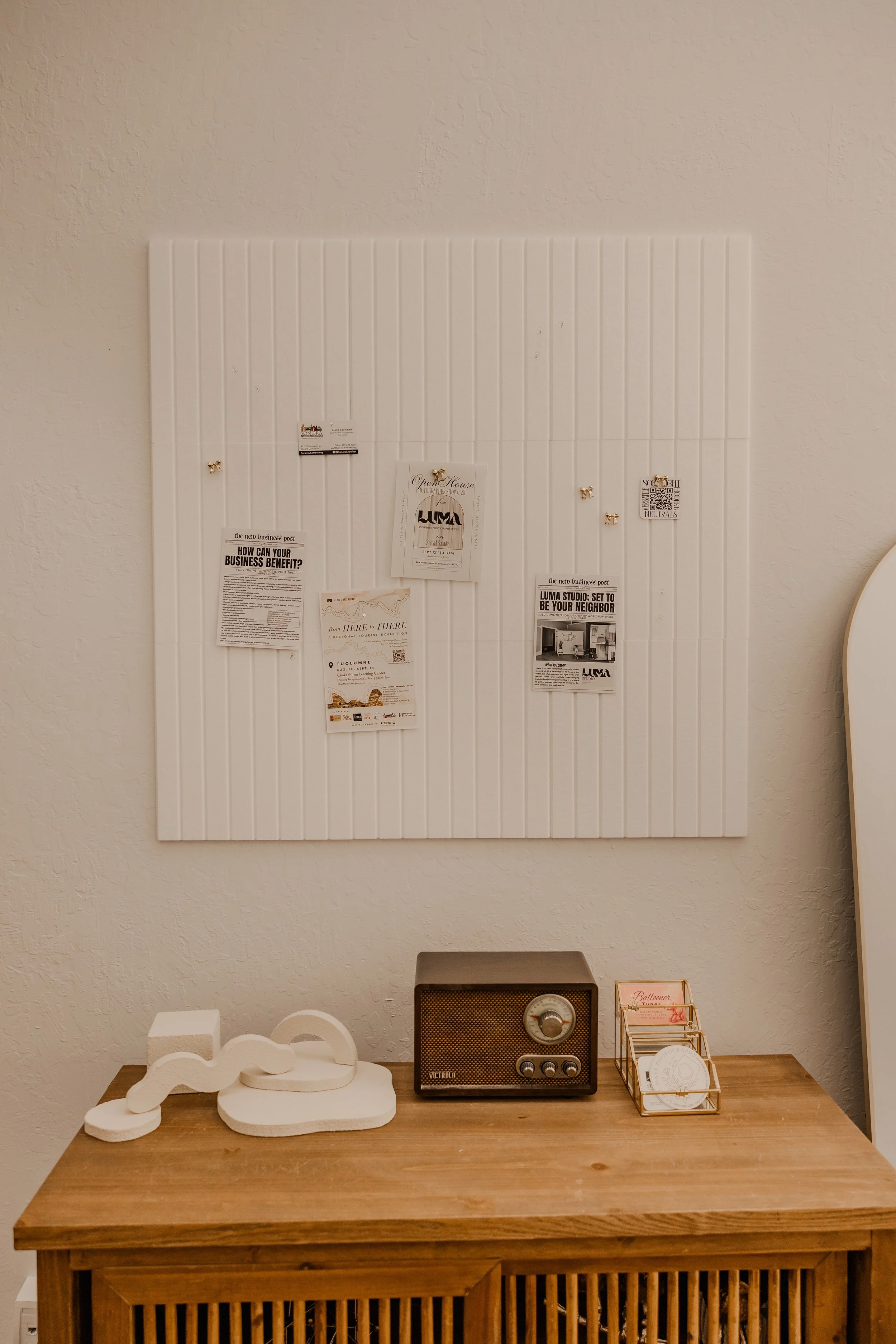 A wooden table with a vintage radio, a small wire basket with cards, and white decorative objects, with a white wall behind displaying flyers and notices.