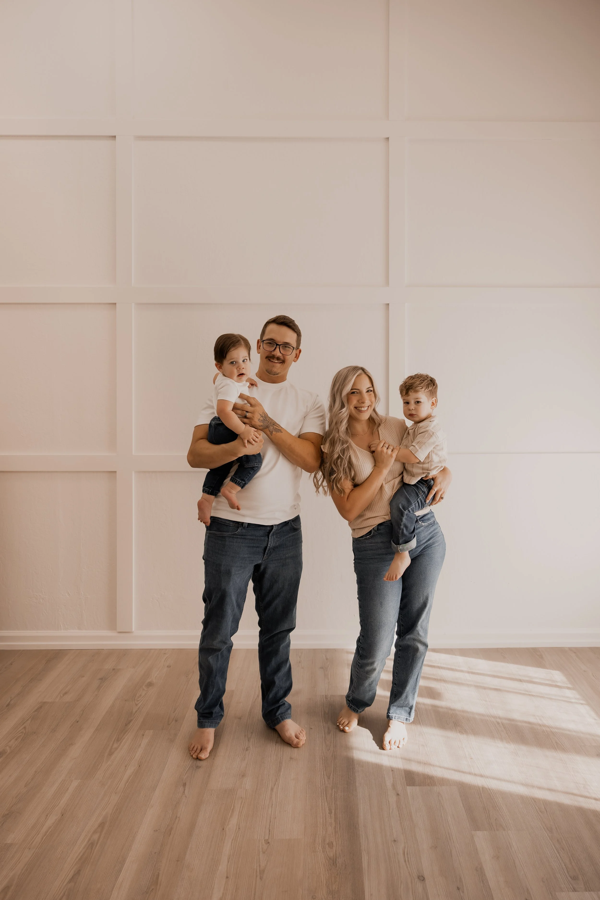 A smiling family of four standing barefoot in a room with wooden floor and white wall, parents holding two children.