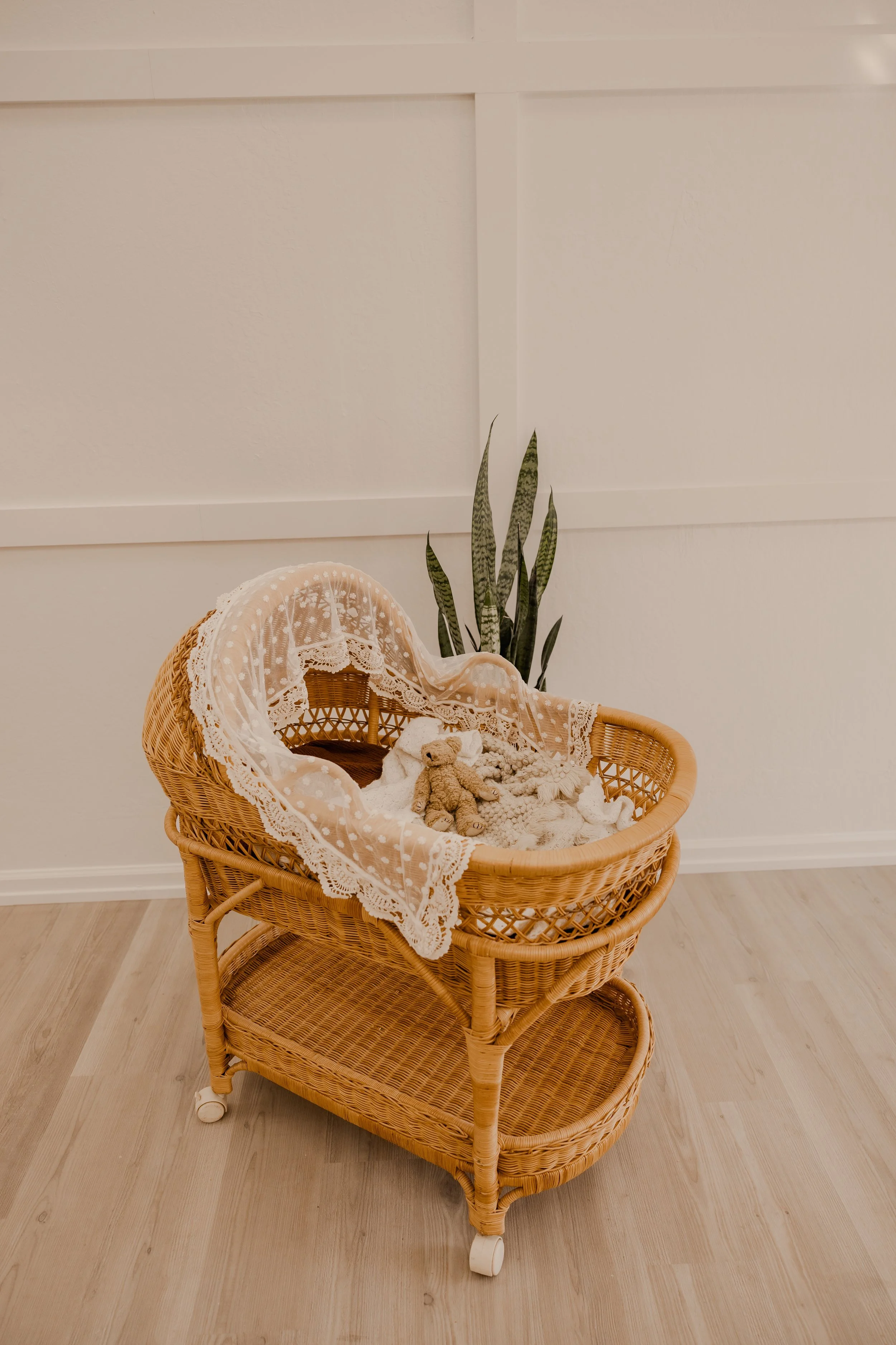 Wicker baby cradle with lace blanket and stuffed animals inside, positioned on a light wood floor against a white wall with panel molding and a tall green plant beside it.