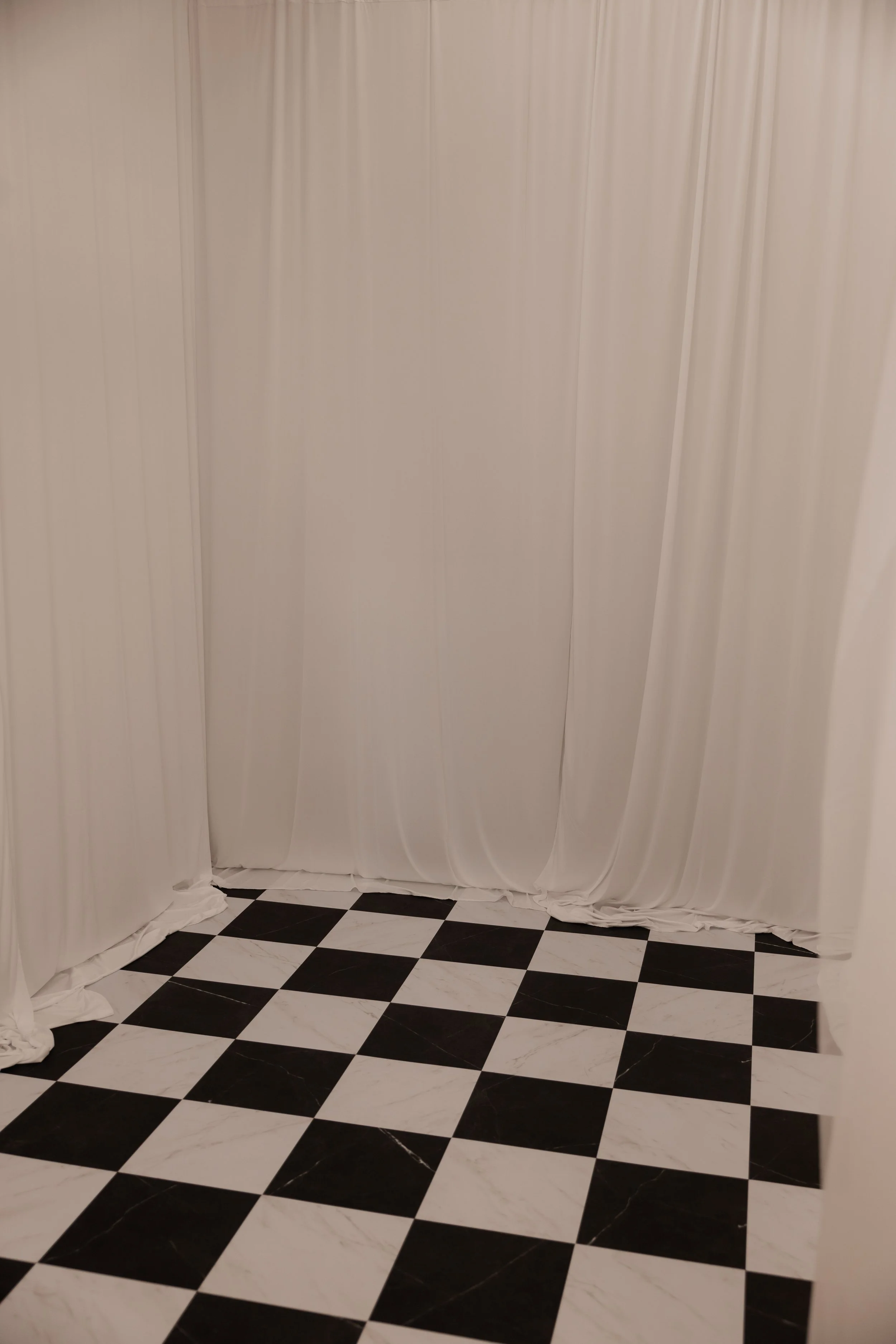 A corner of a room with a black and white checkered floor and white curtains hanging from ceiling to floor.