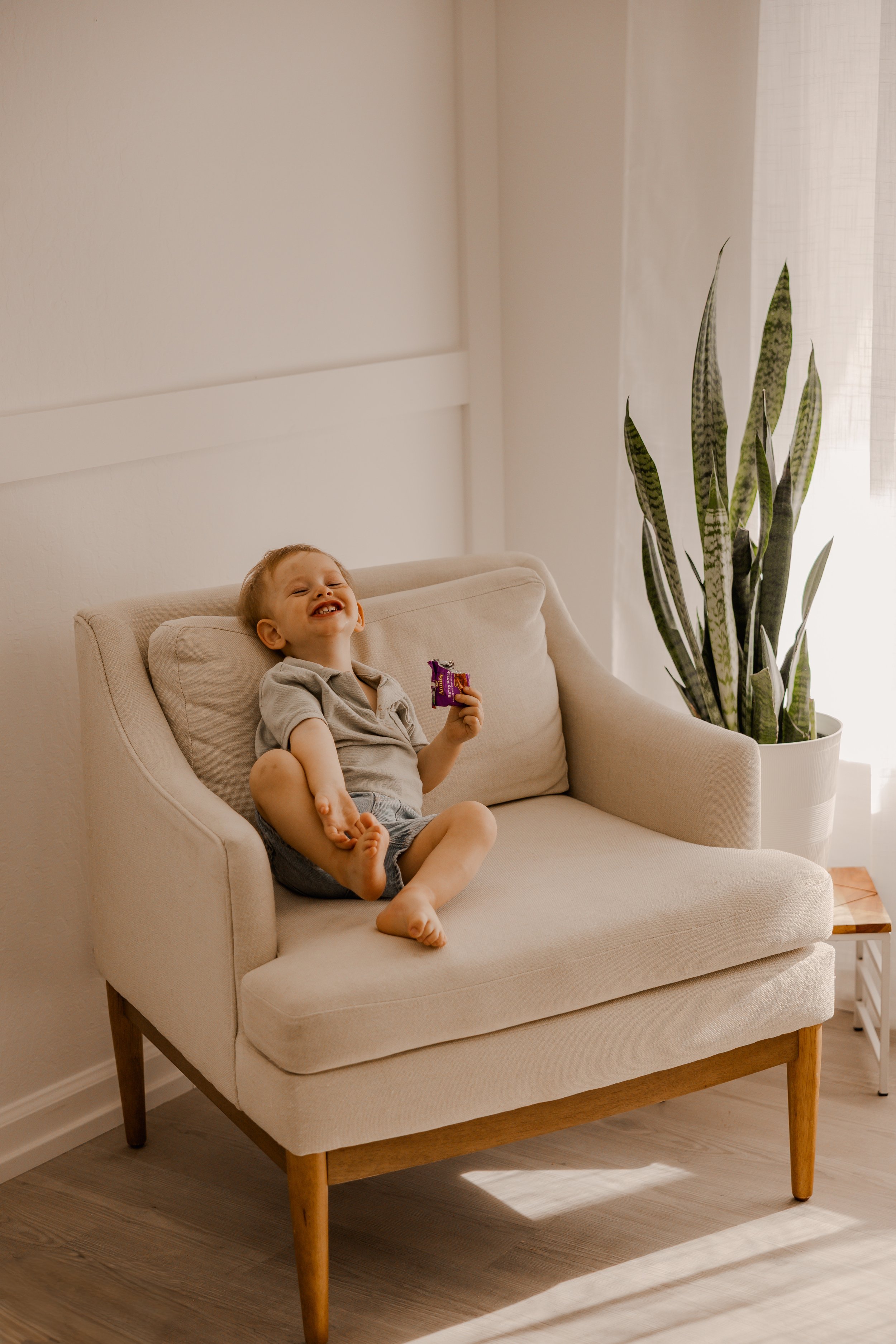 A young boy sitting on a beige armchair, smiling and holding a small snack, with a large green houseplant nearby in a bright room.