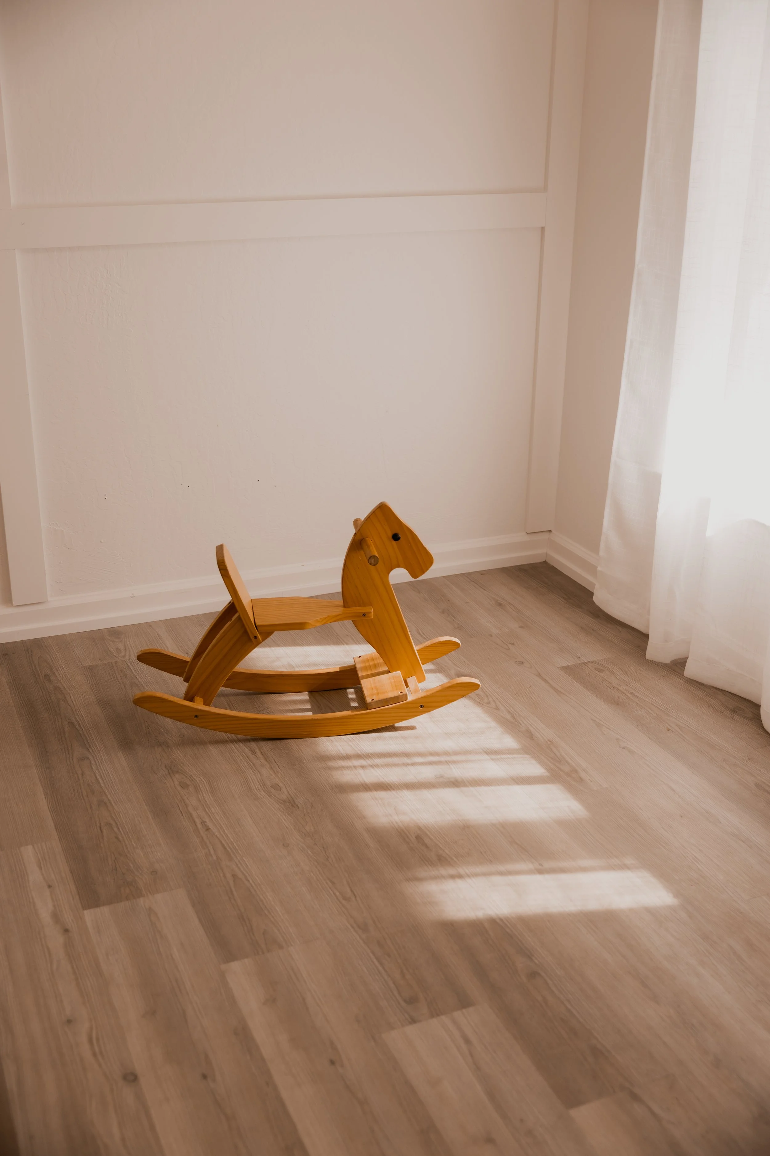 A wooden rocking horse toy on a light-colored wooden floor near a white wall and a white curtain with sunlight streaming through.