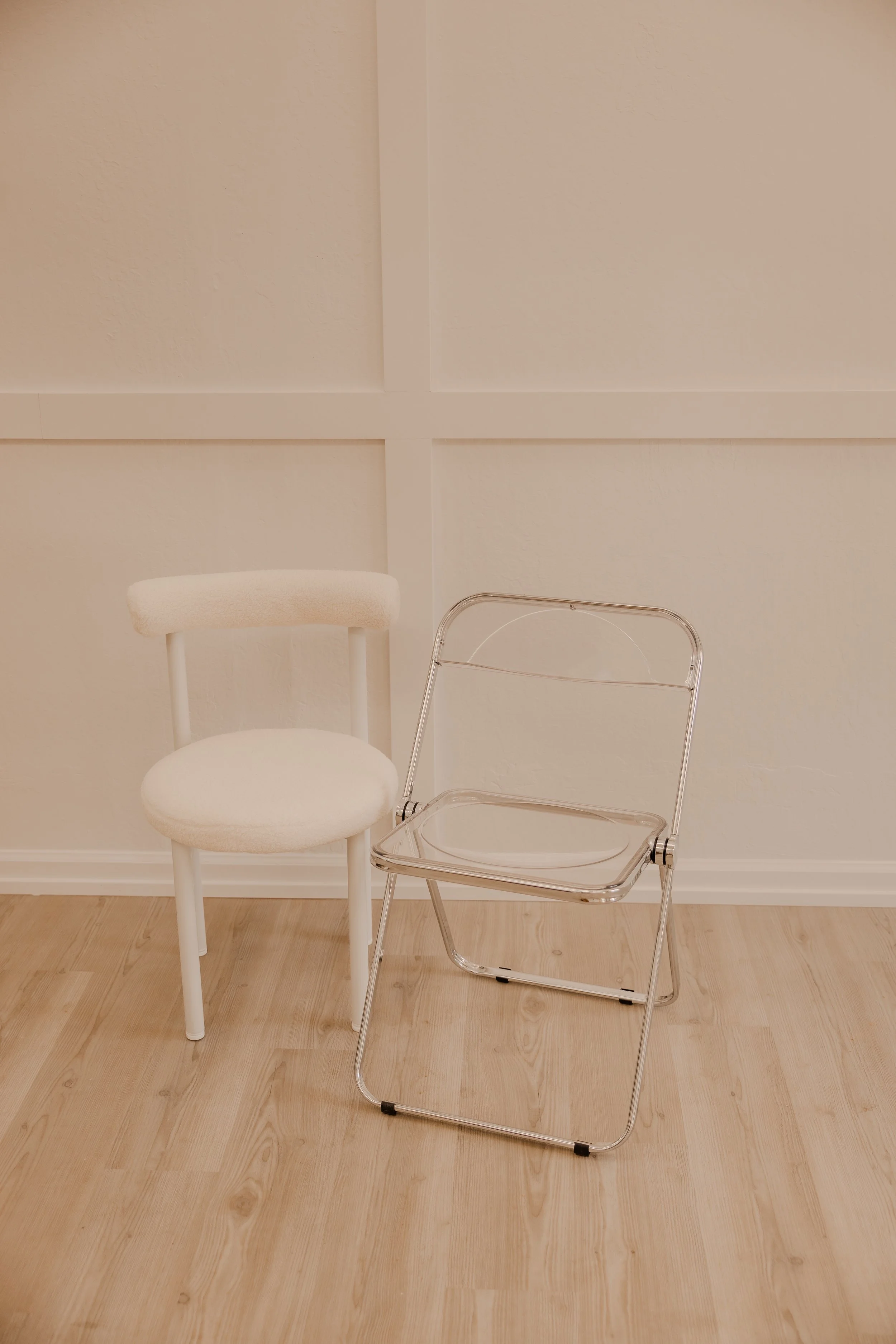 A white chair with a fluffy backrest and seat next to a clear plastic chair with a metal frame, set against a beige wall with wooden flooring.