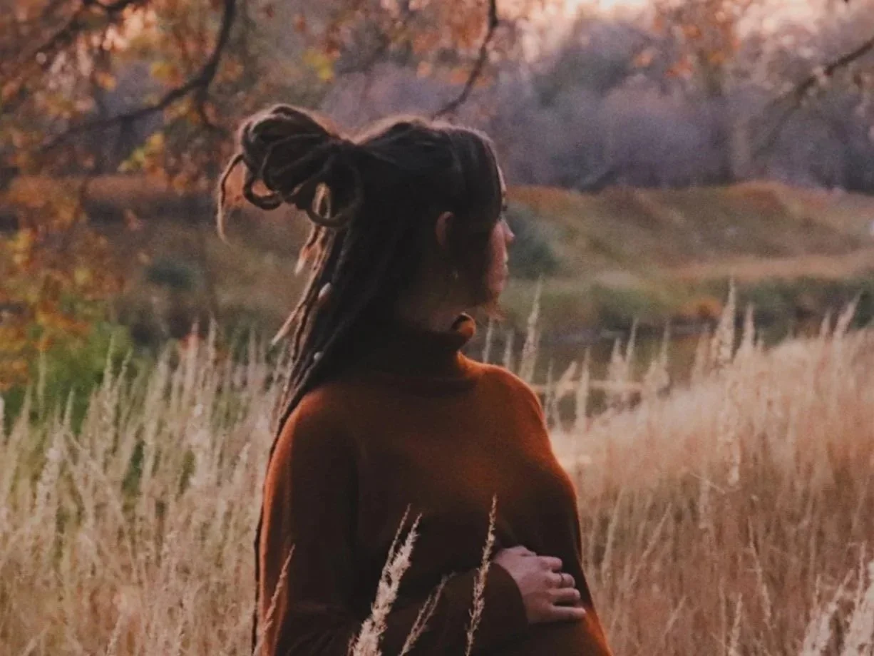 A person with dreadlocks standing in a field of tall grass during sunset, with trees and a lake in the background.
