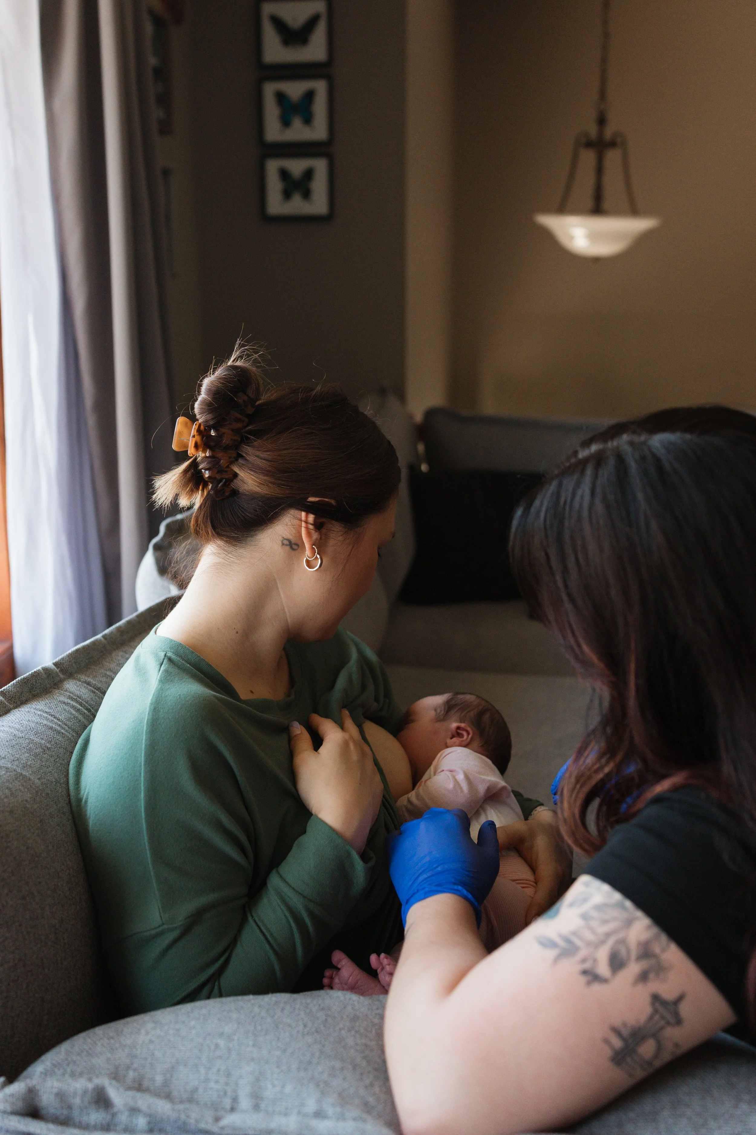 A woman with a hair bun and earrings breastfeeding a baby while another woman with tattoos and a leg cast interacts with them in a cozy living room.