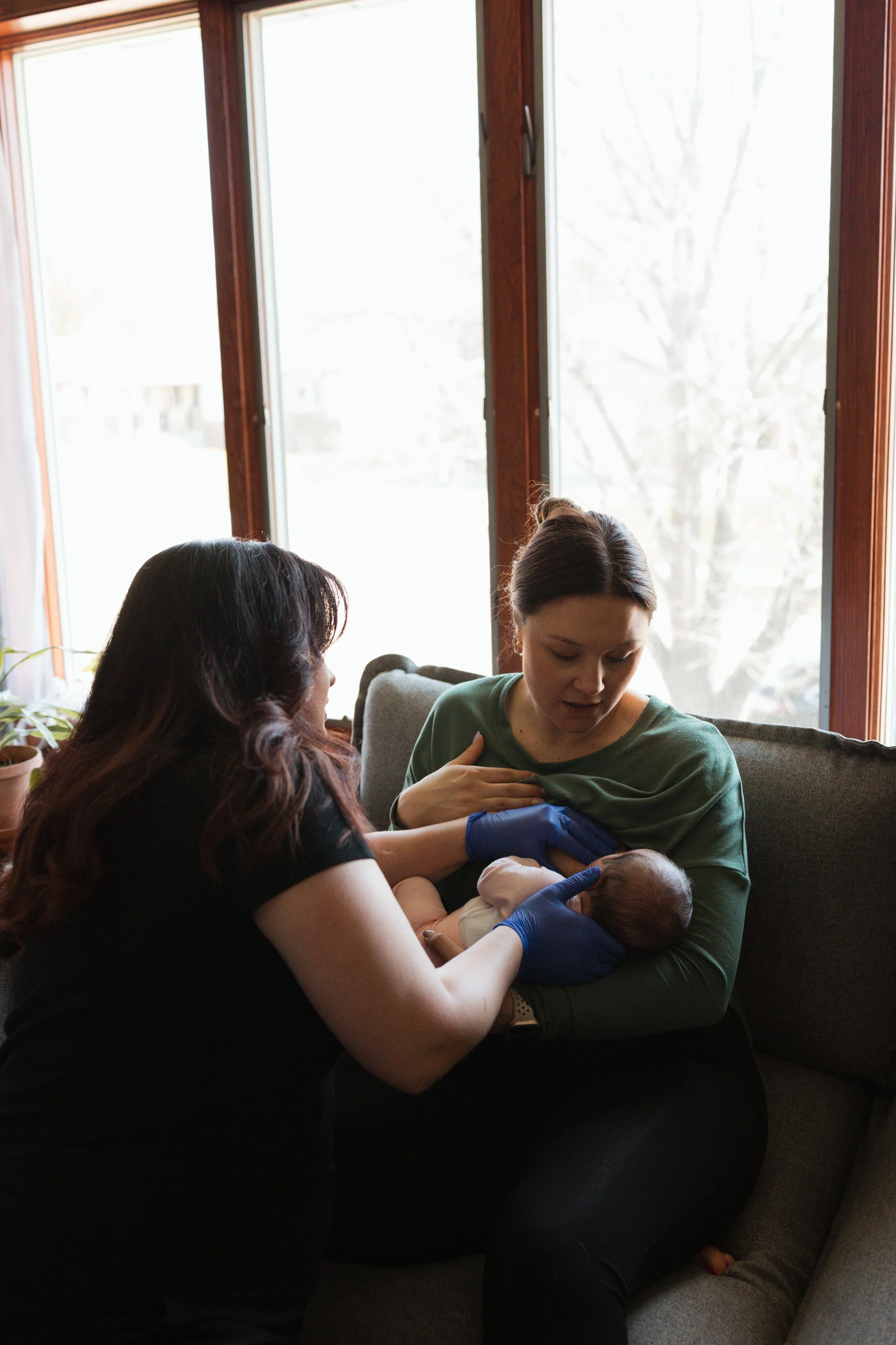 A woman in a green shirt holds a newborn baby while sitting on a gray couch next to another woman with dark hair. The second woman is helping with the baby, wearing purple gloves. They are inside a room with large windows showing a tree outside.