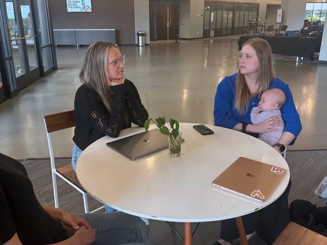 Two women sitting at a round table in a lobby or waiting area, one holding a baby, with laptops and a phone on the table.