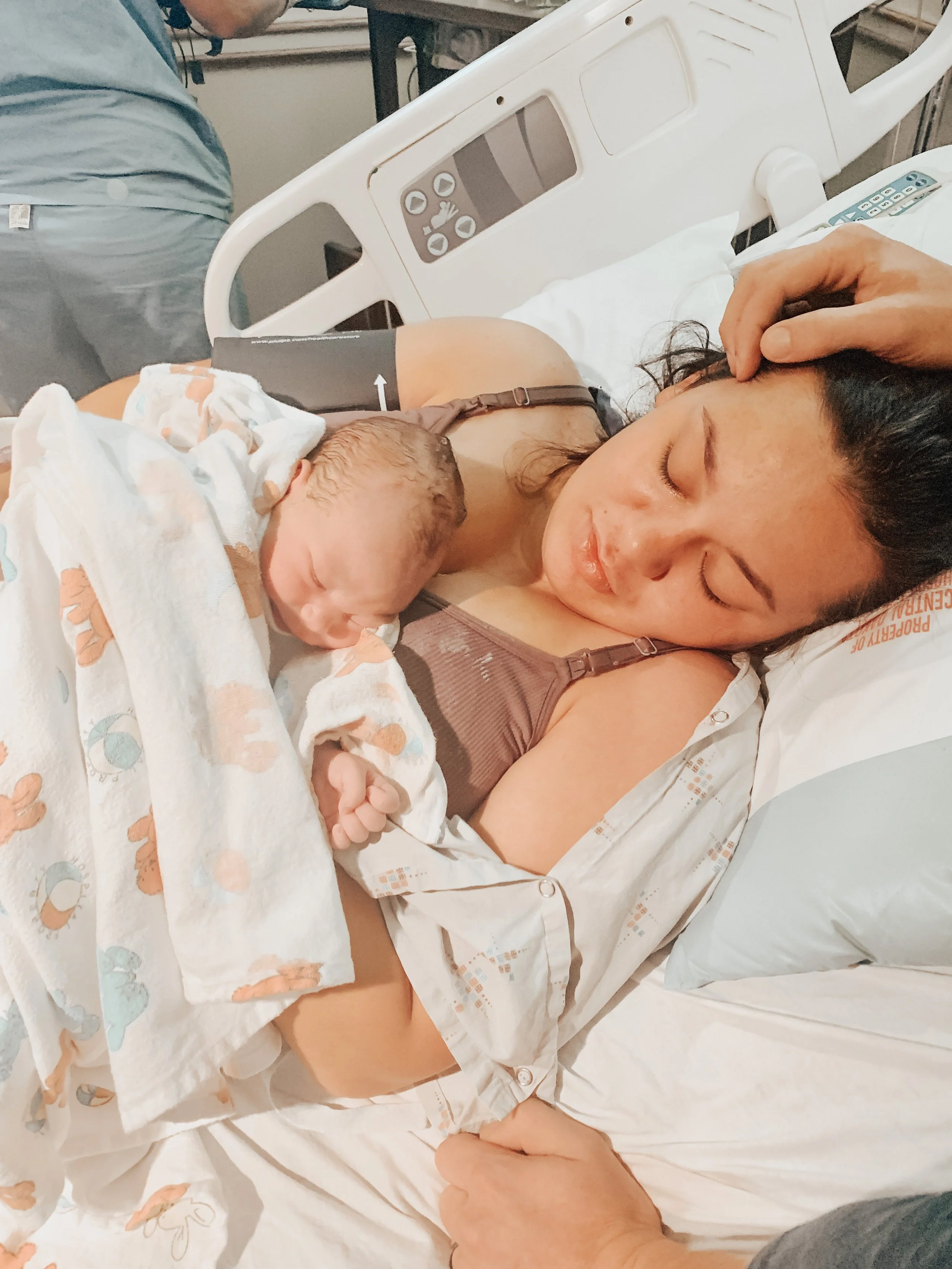 A woman is lying in a hospital bed, holding her newborn baby close to her chest. The woman appears to be resting with her eyes closed, and the baby is wrapped in a hospital blanket. A hand is gently touching her forehead. Medical equipment and a person in scrubs are visible in the background.