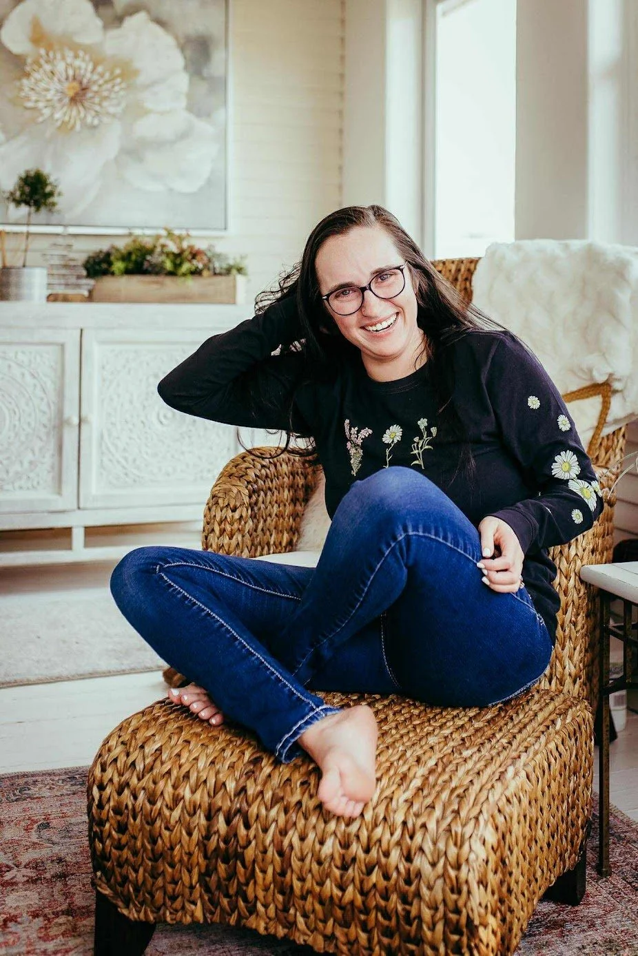 A woman with glasses and long dark hair sitting on a wicker chair in a bright room, smiling, wearing a black sweater with daisies and blue jeans.