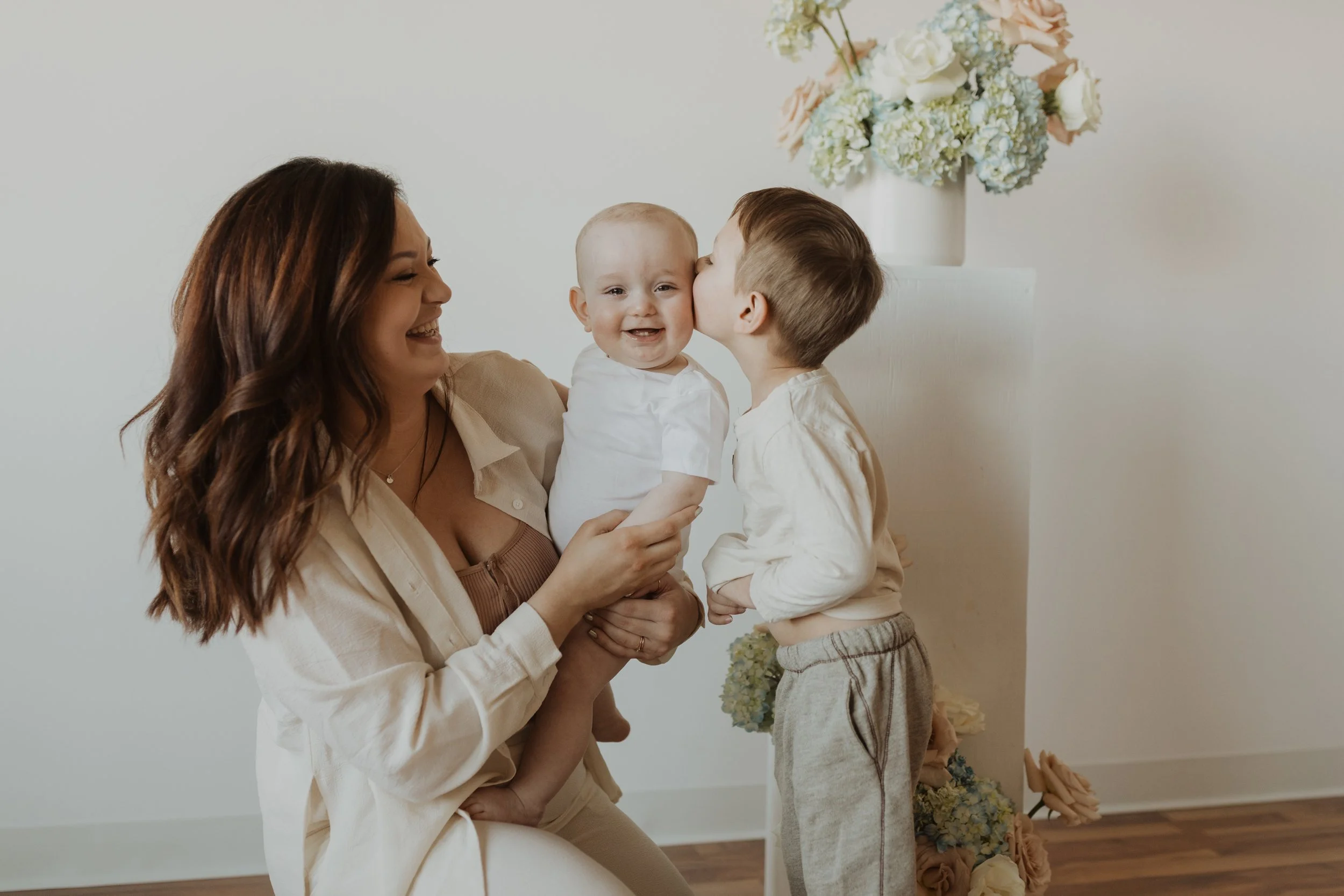 A woman holding a smiling baby while a young boy kisses the baby's cheek, in a room with white walls and a floral arrangement in the background.