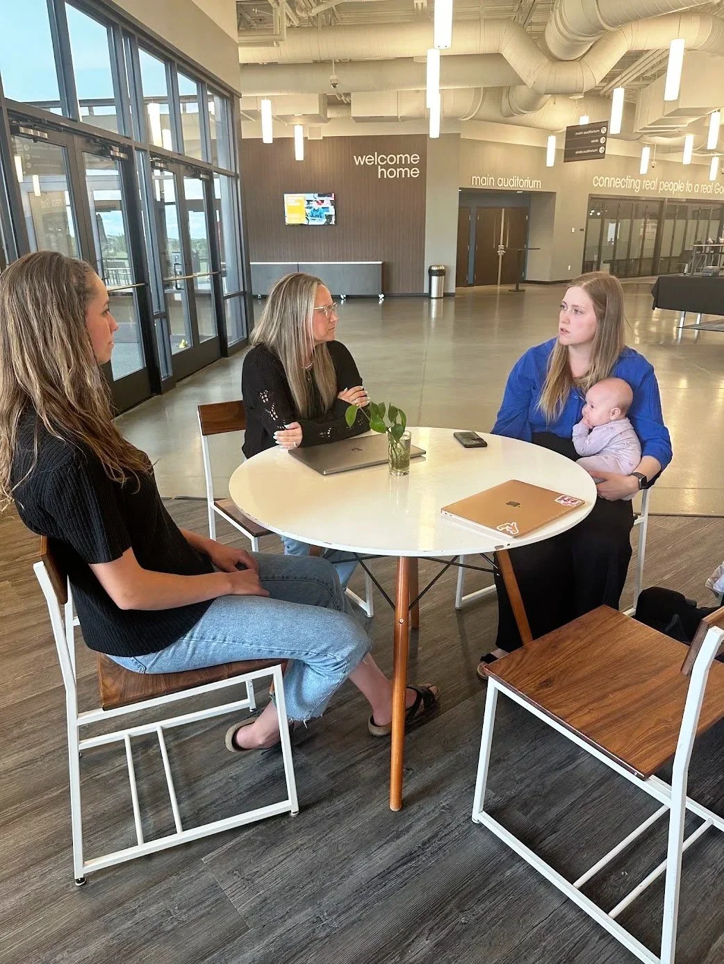 Four women sitting around a round table in an open lobby area, one woman holding a baby, other women with laptops and phones, large windows in the background with signs reading 'welcome home,' 'main auditorium,' and 'connecting real people to a real God.'