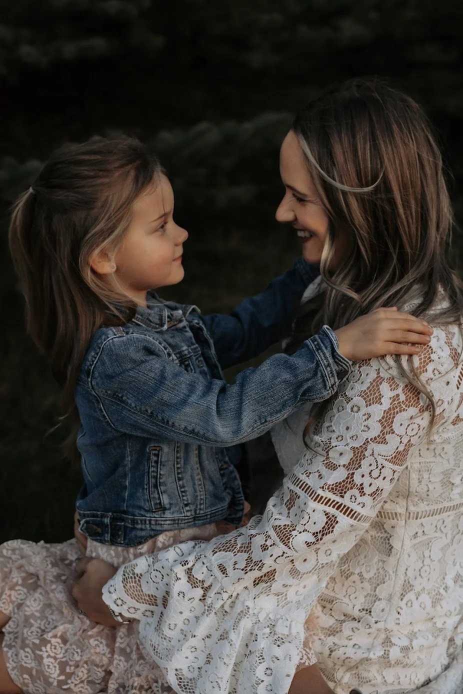 A woman and a young girl sitting outdoors, smiling and facing each other. The girl is wearing a denim jacket and the woman is dressed in a white, lace dress.