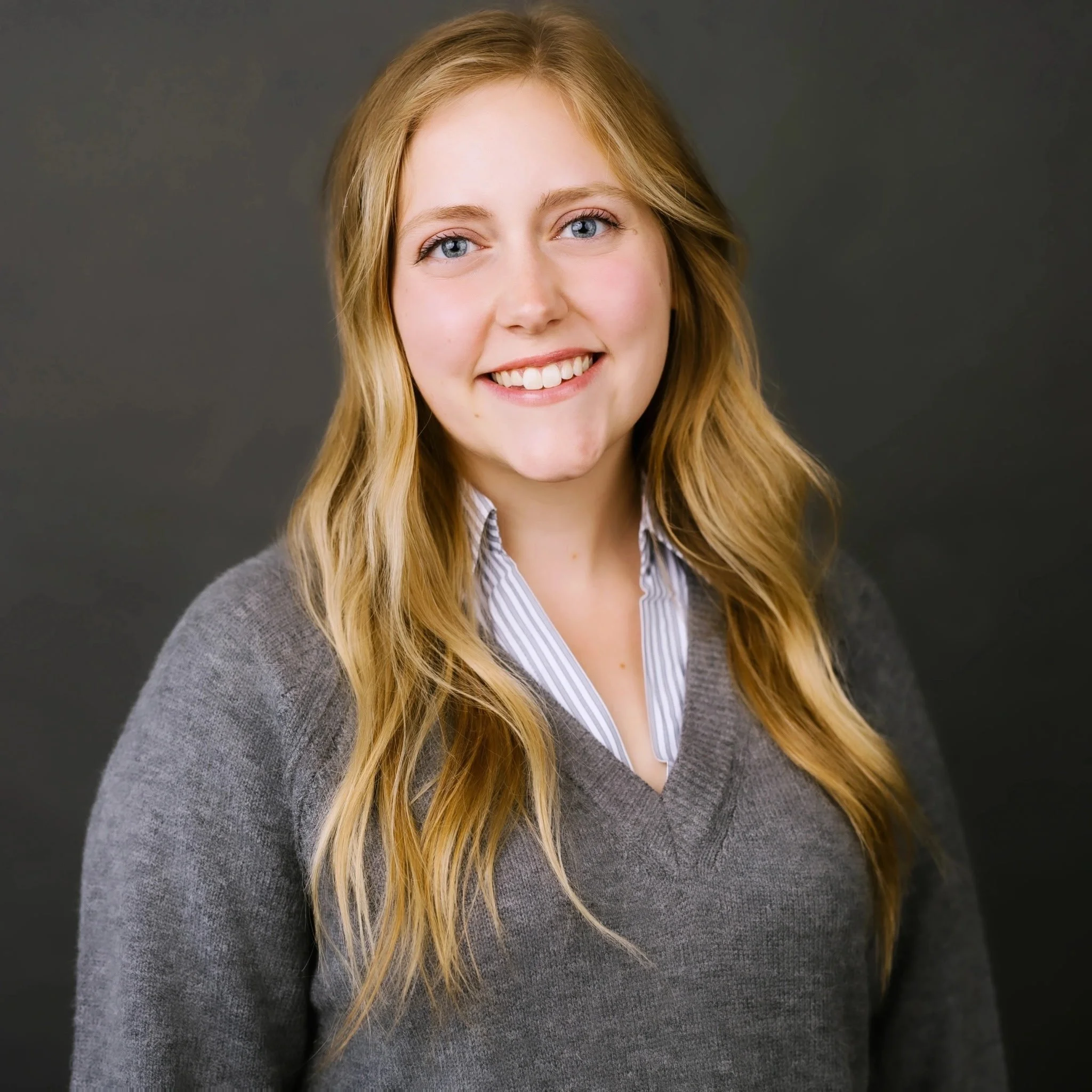 Close-up of a young woman with long blonde hair, blue eyes, smiling, wearing a gray sweater over a striped collared shirt, against a dark background.