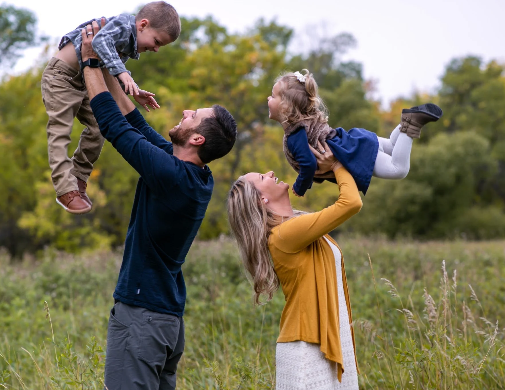 A family of four outdoors in a grassy field with trees, with the mother, father, and daughter lifting the son in the air, laughing and smiling.