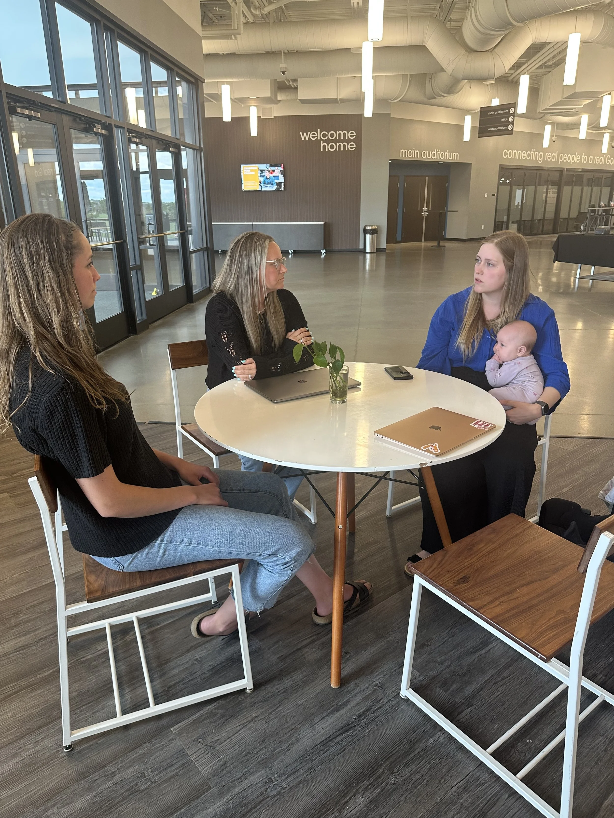 Four women and a baby sitting around a round table in a modern indoor space with large glass doors, a TV, and signage that includes phrases like 'welcome home' and 'main auditorium'.