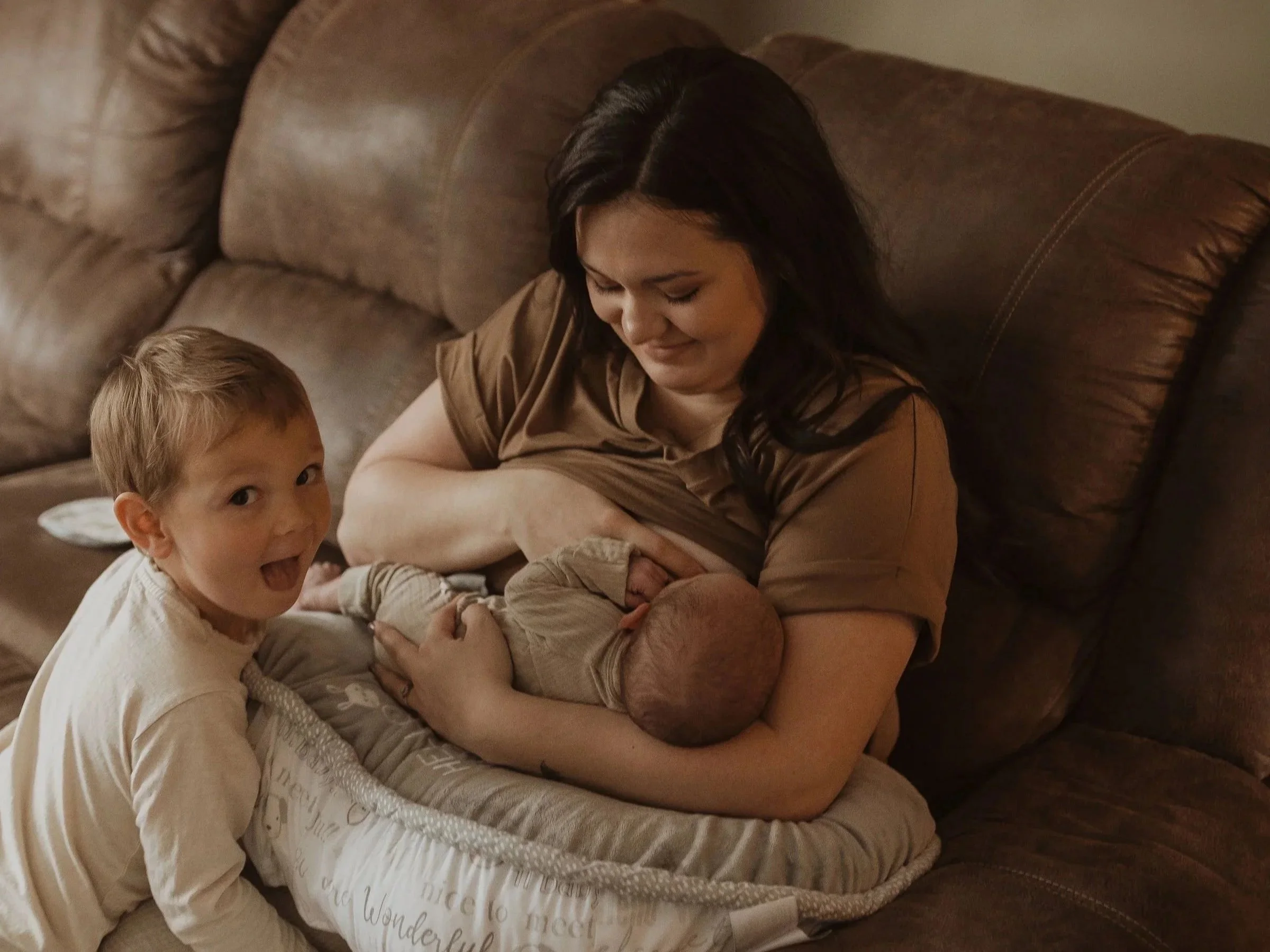 A woman with dark hair smiling down at a young boy with light brown hair, both sitting on a brown leather couch. The boy is looking at the camera with an open-mouthed smile.