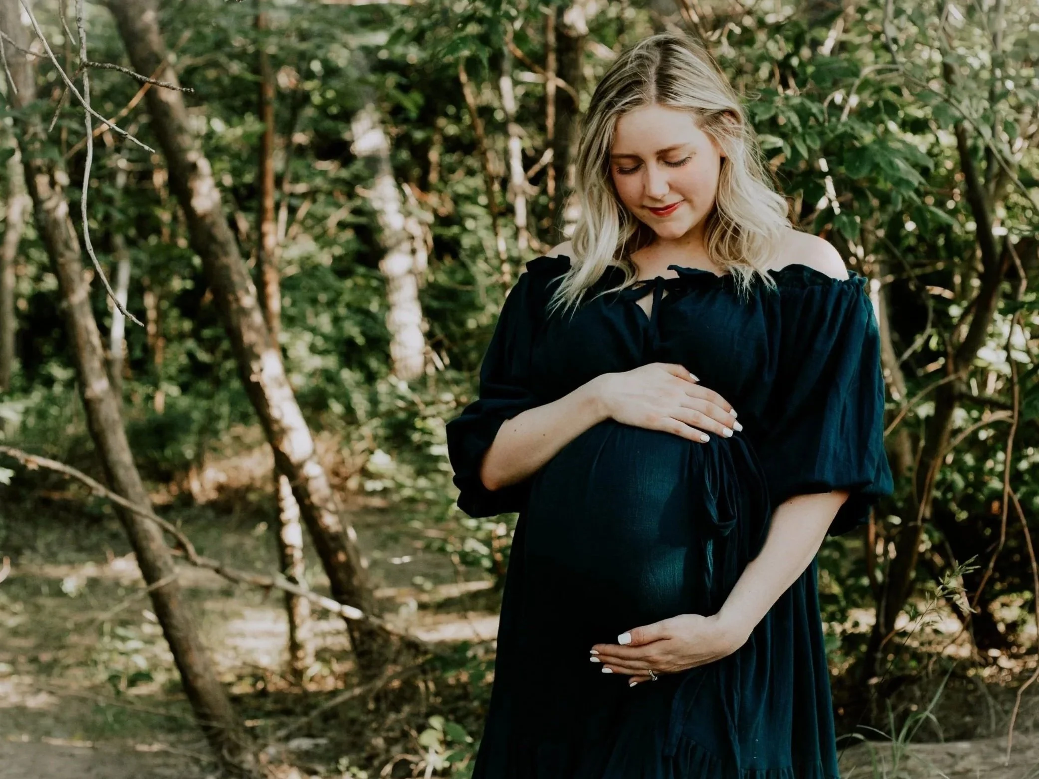 A pregnant woman with blonde, wavy hair wearing a dark off-the-shoulder dress standing outdoors among trees, gently resting her hands on her stomach and looking down with a serene expression.