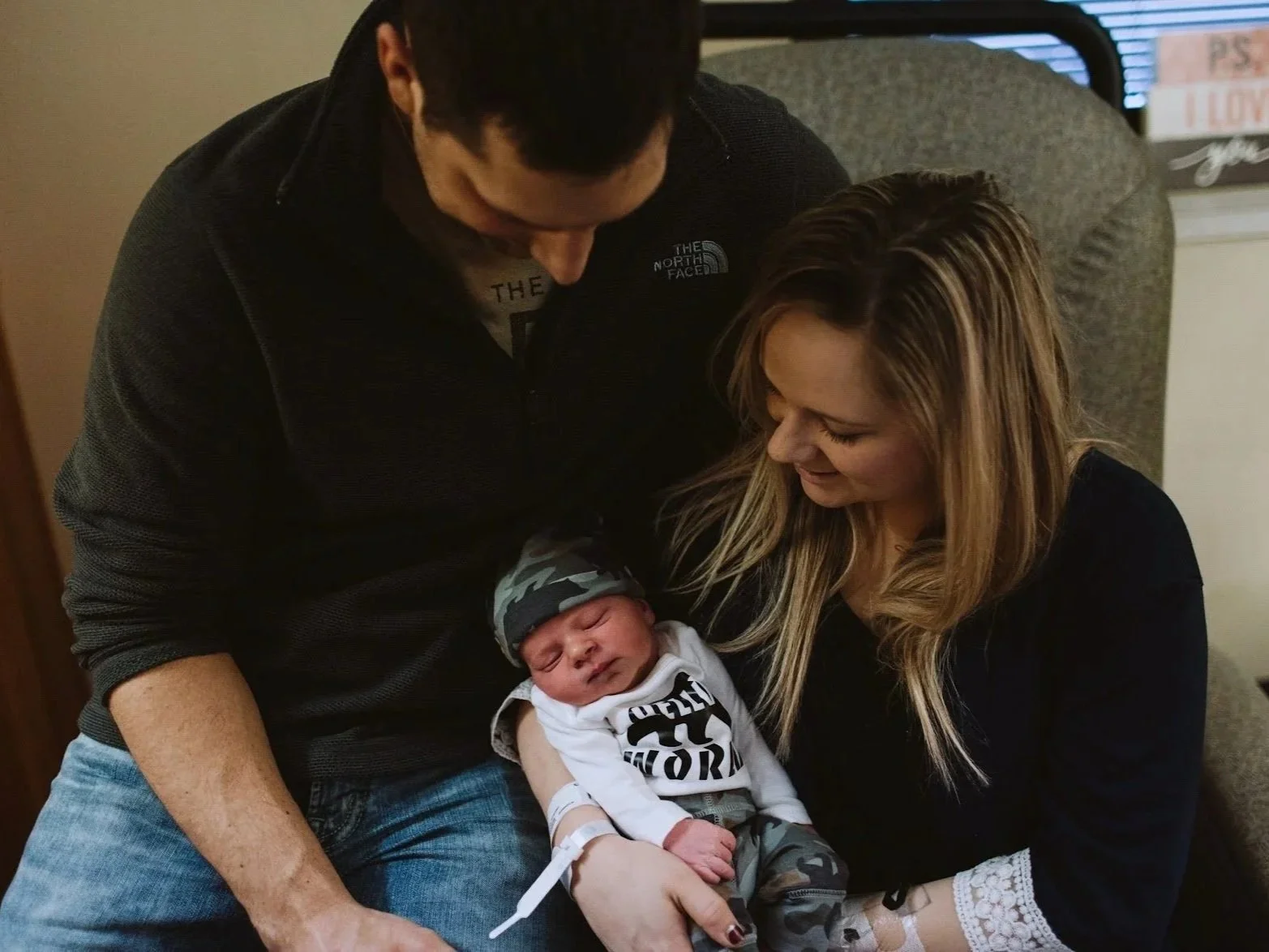 A couple holding a newborn baby, with the man looking down at the baby and the woman smiling at the baby. The baby is wrapped in a blanket and wearing a hat.