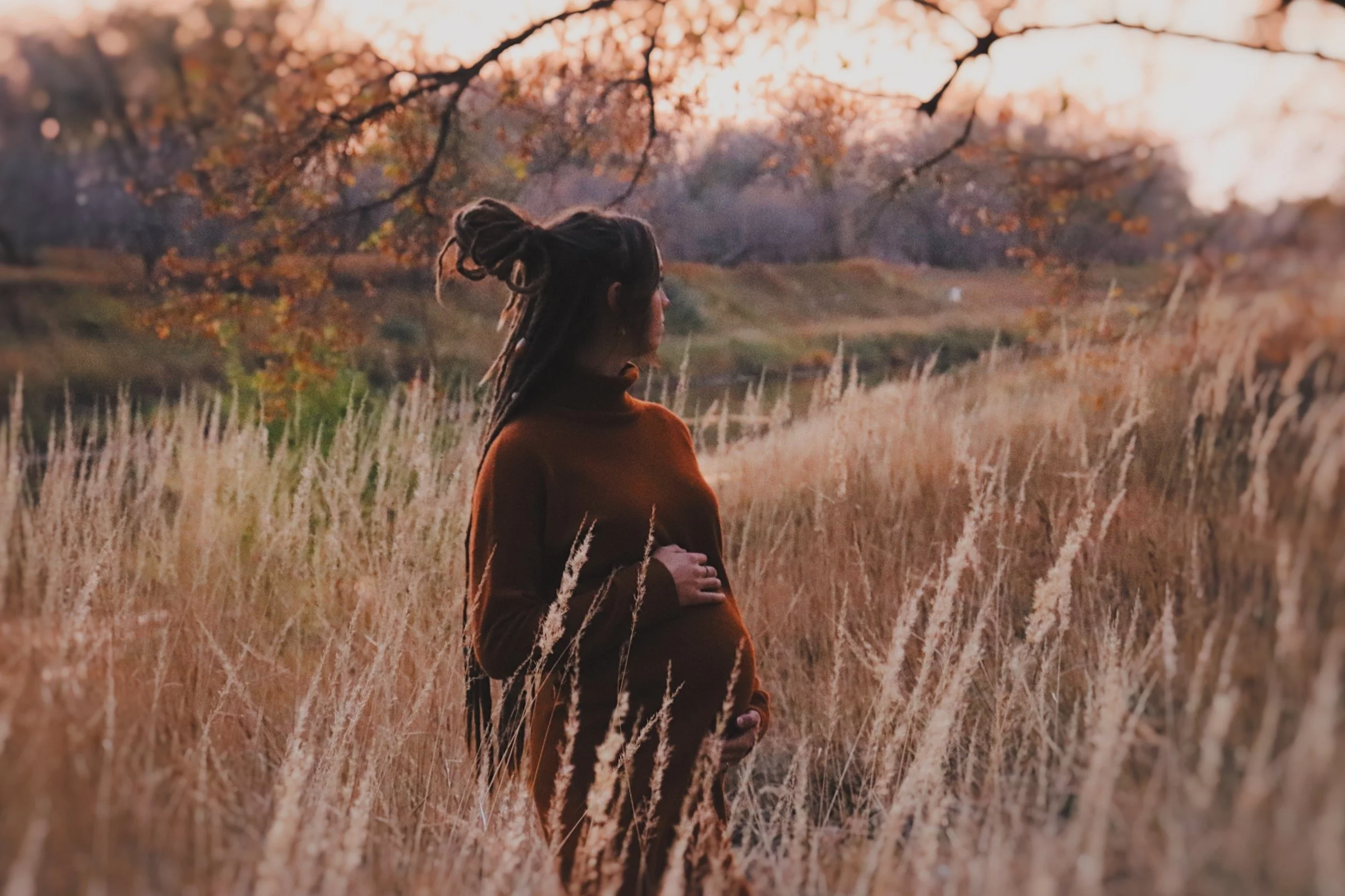 A woman with dreadlocks wearing a brown jacket standing in a field of tall grass with trees and an open landscape in the background during sunset.